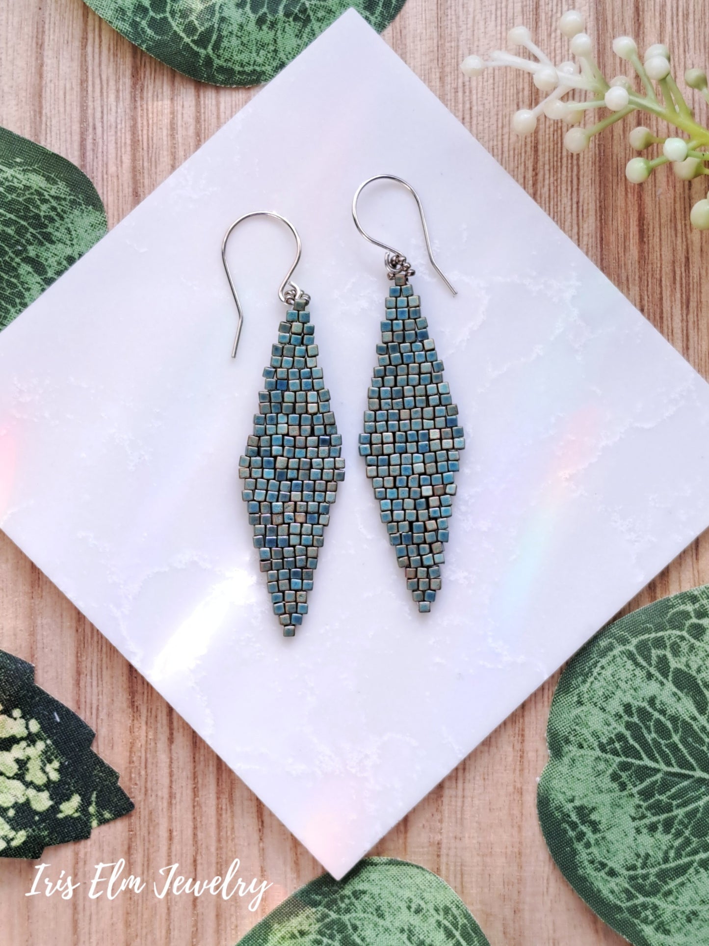Pair of beaded turquoise glass matte beadwork diamond shape earrings on a white square with green leaves around