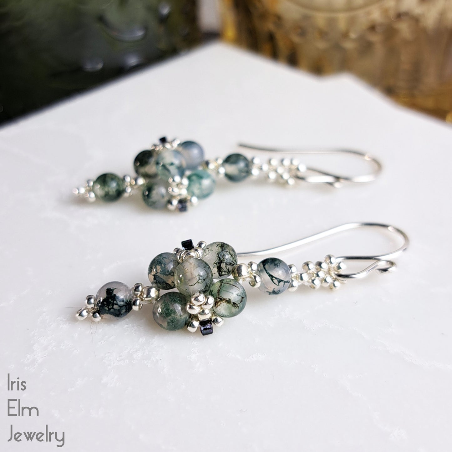 Pair of silver beaded earrings with green moss agate gemstones on a white surface, branded 'Iris Elm Jewelry'.