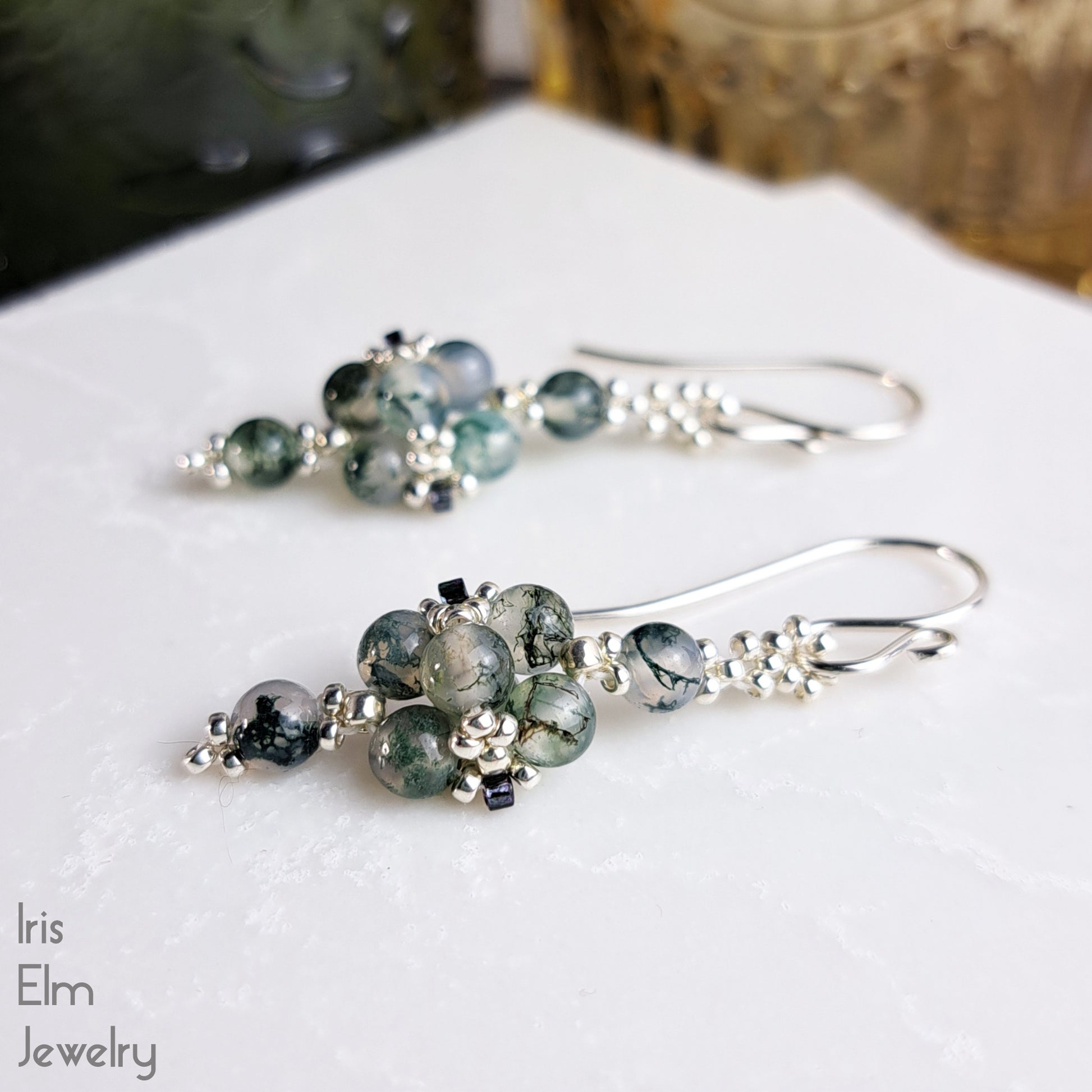 Pair of silver beaded earrings with green moss agate gemstones on a white surface, branded 'Iris Elm Jewelry'.