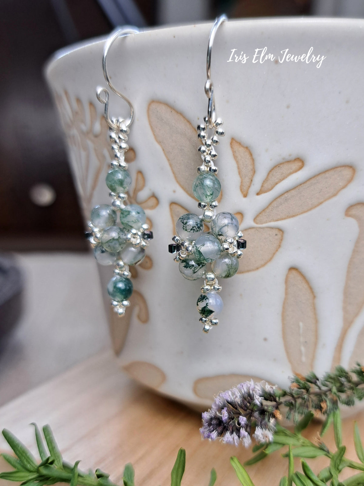 A pair of silver dangle earrings with green moss agate beads, hanging from a ceramic mug. The image has a watermark 'Iris Elm Jewelry' and features decorative rosemary and lavender sprigs in the foreground.