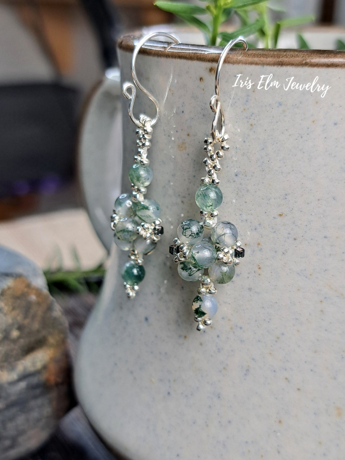 A pair of silver dangle earrings with green marbled moss agate beads and small silver accents, hanging from a speckled gray ceramic mug. The brand name 'Iris Elm Jewelry' is watermarked on the upper right