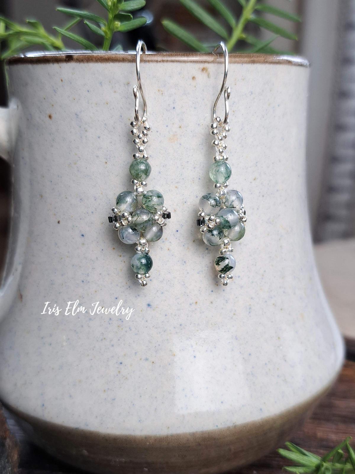 A pair of silver dangle earrings with green moss agate beads and silver seed beads, displayed on a speckled ceramic mug with green foliage in the background. The brand name 'Iris Elm Jewelry' is watermarked on the lower left.