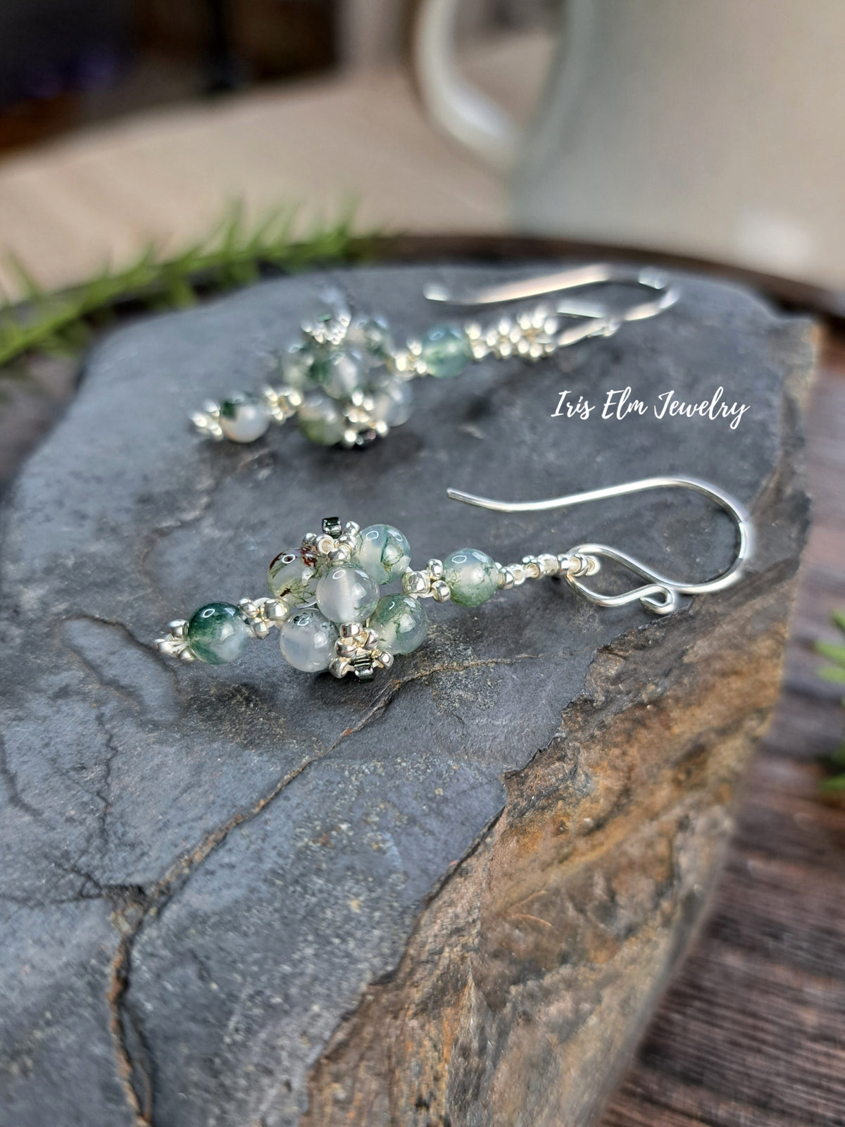 A pair of handmade dangle earrings by Iris Elm Jewelry, featuring silver-toned metal hooks and clusters of green moss agate beads, displayed on a dark textured stone surface.