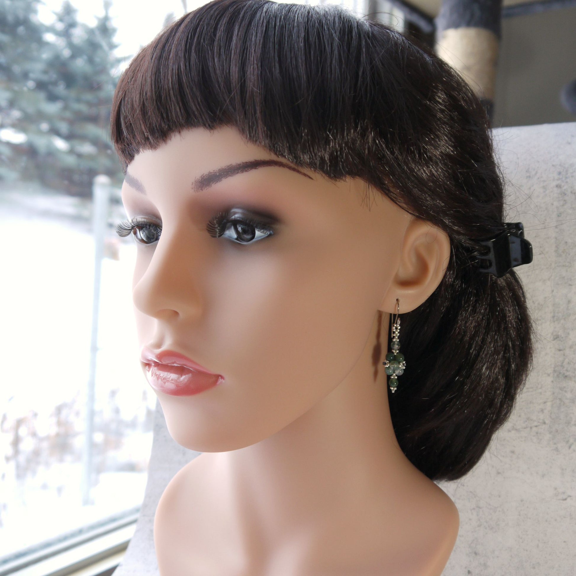 Mannequin head with a black wig and small moss agate earrings on a blurred background