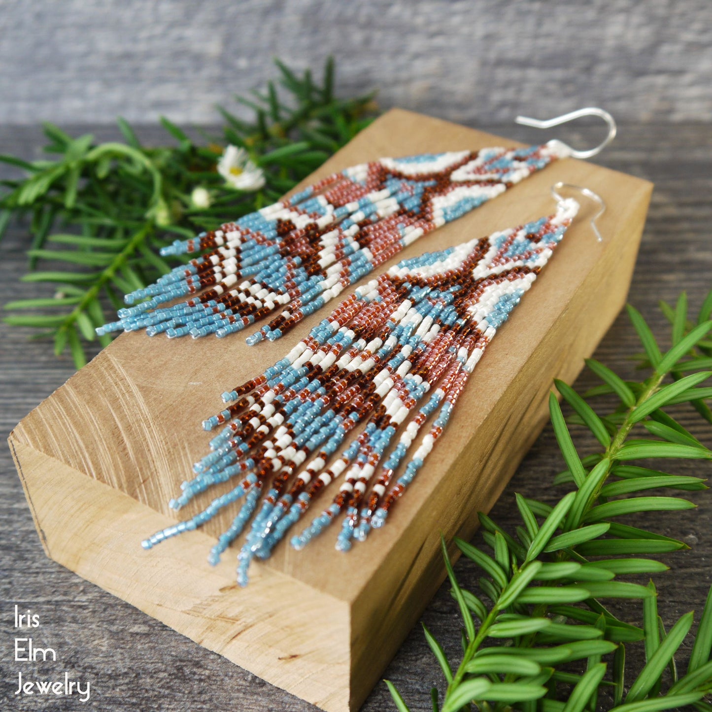 Beaded glass seed bead brown and blue fringe geometric earrings on a wooden block with greenery around