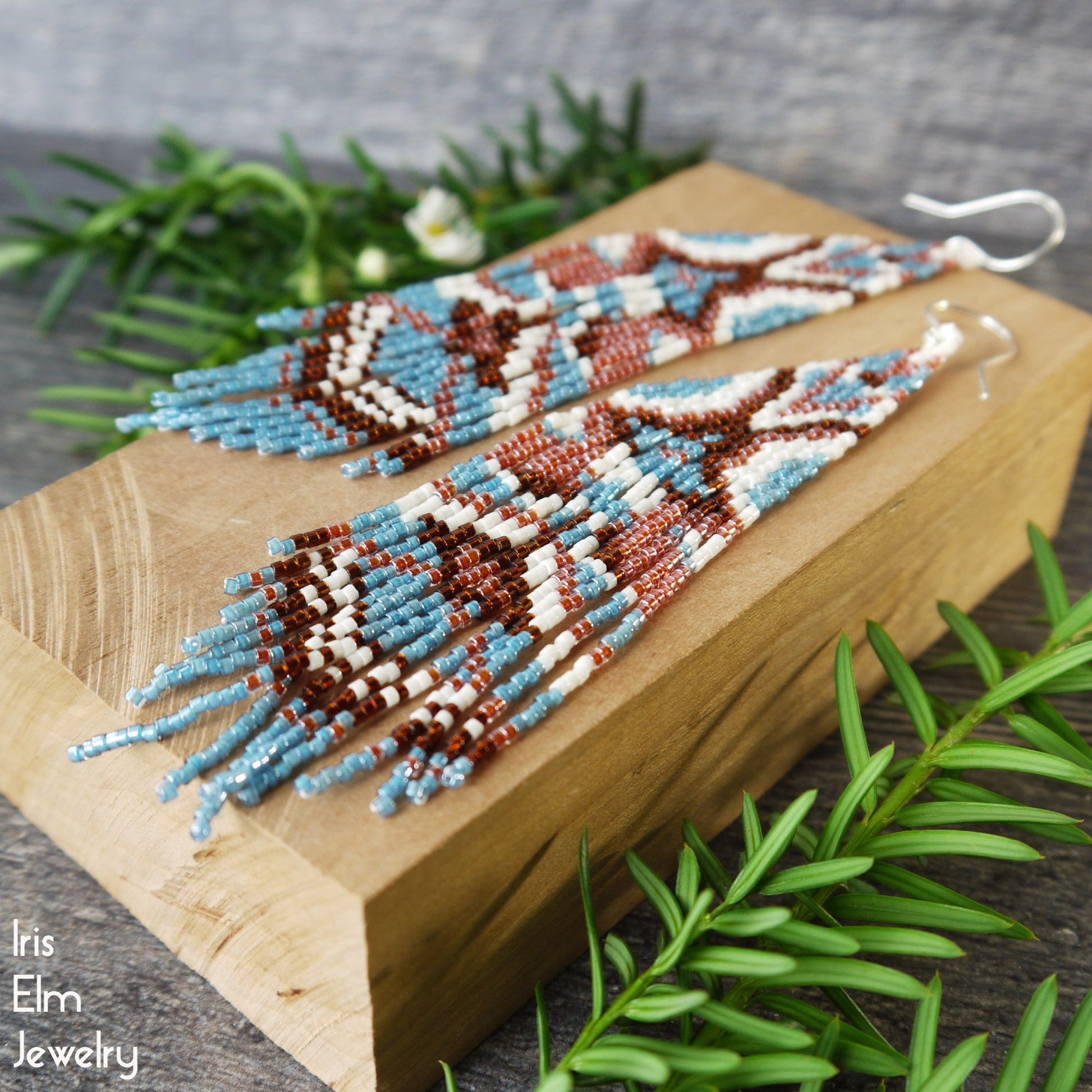 handmade seed bead long fringe blue, white, orange, and brown earrings on a wooden block with greenery around