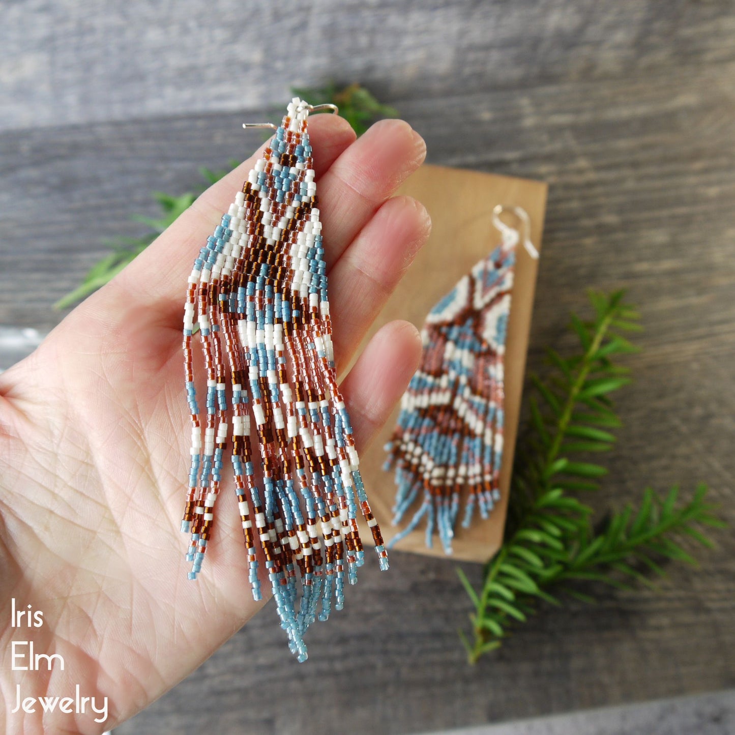 Hand holding beaded seed bead fringe earrings with a wooden display card and greenery in the background, featuring 'Iris Elm Jewelry'.