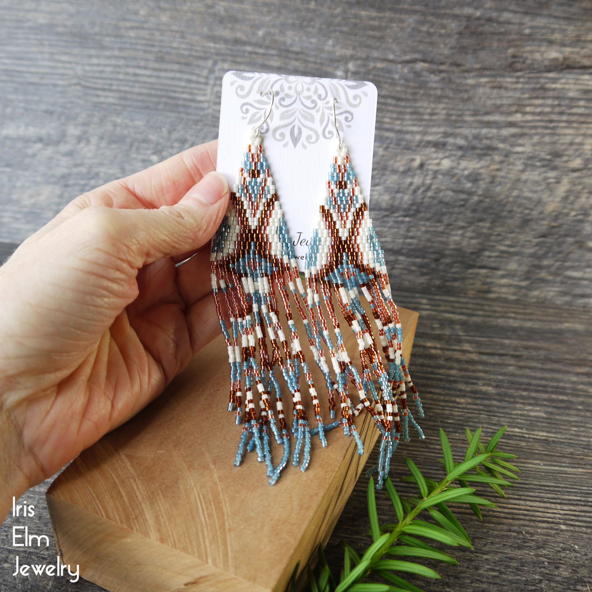 Hand holding beaded geometric seed bead fringe earrings on a wooden surface with 'Iris Elm Jewelry' branding.