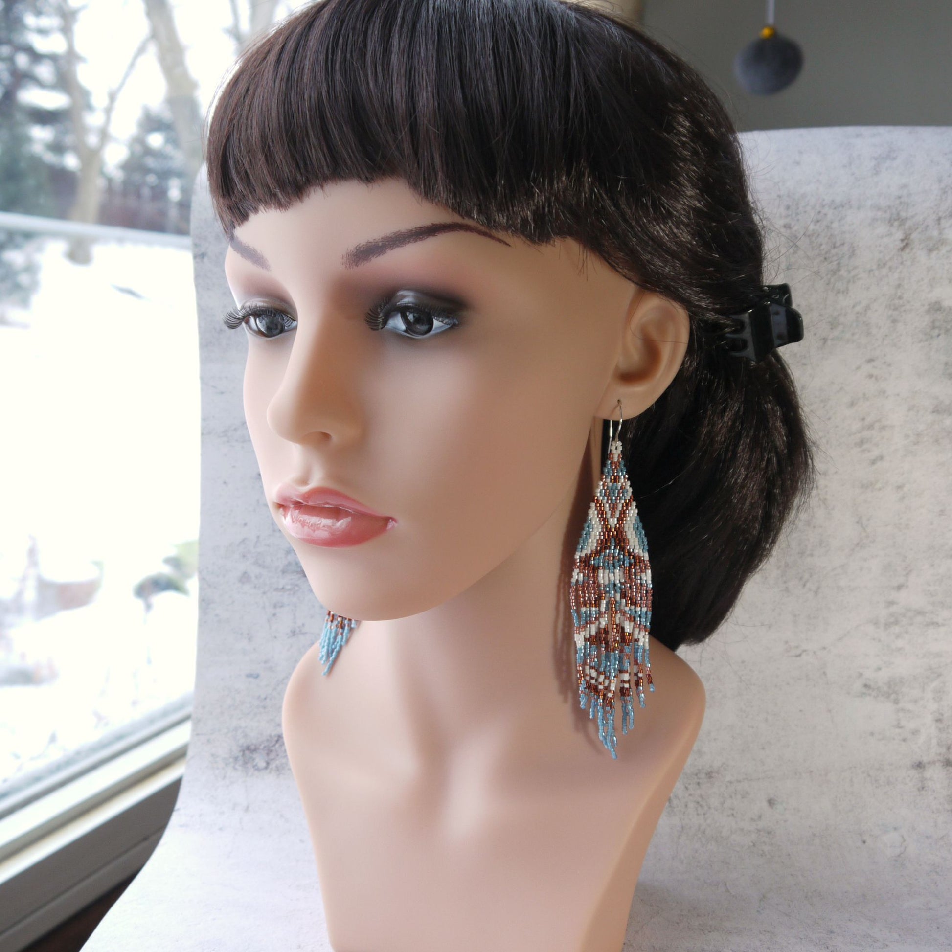 Mannequin head with dark hair wearing long beaded fringe earrings in a blurred indoor setting