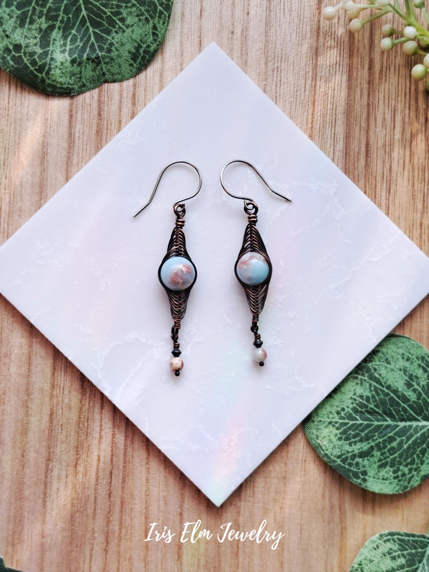 Pair of copper wire wrapped herringbone impression jasper earrings on a white napkin with green leaves on a wooden surface, featuring 'Iris Elm Jewelry'.