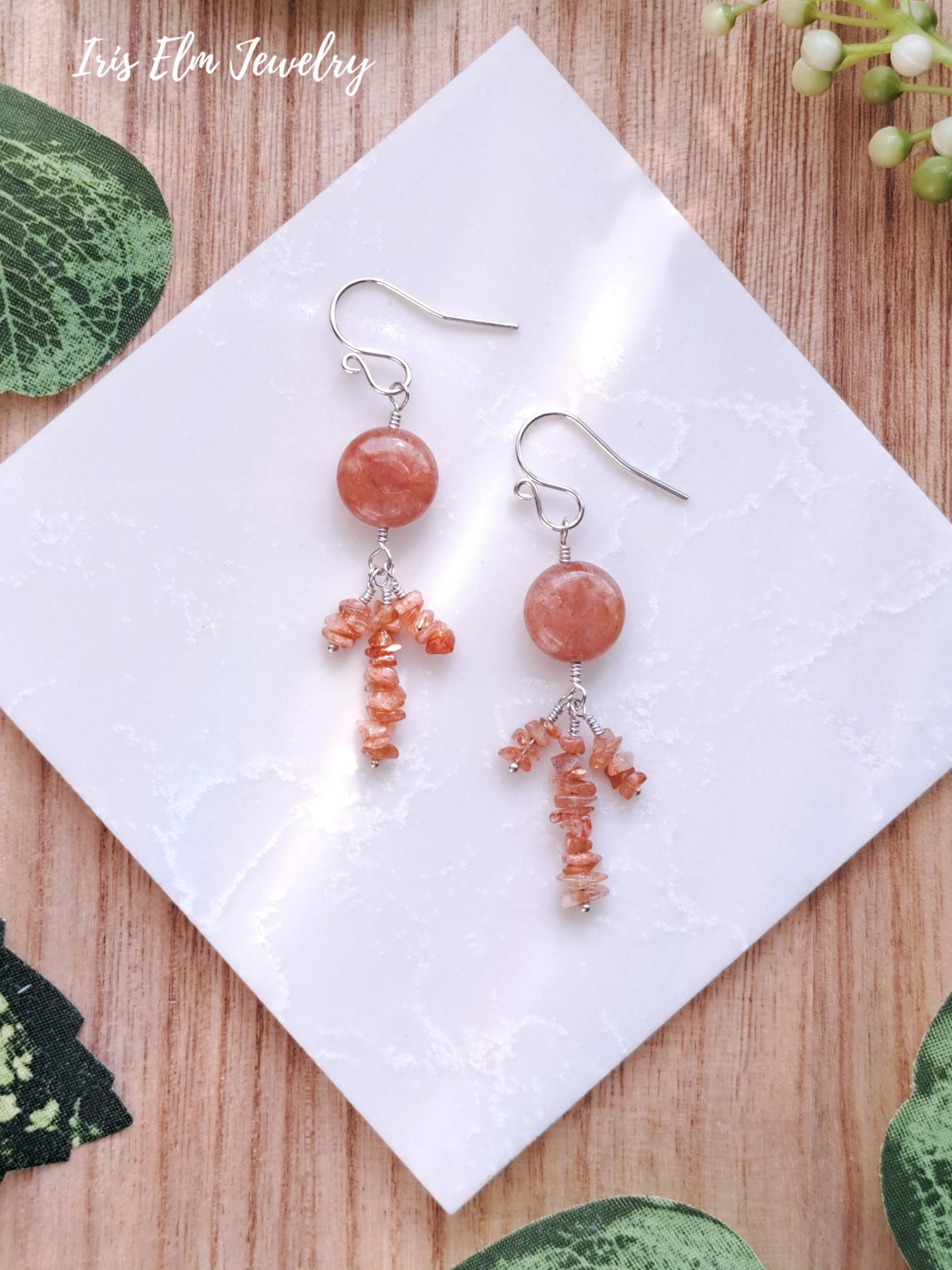 Pair of sunstone and peach agate sterling silver earrings with red beads on a white tile with 'Iris Elm Jewelry' branding.
