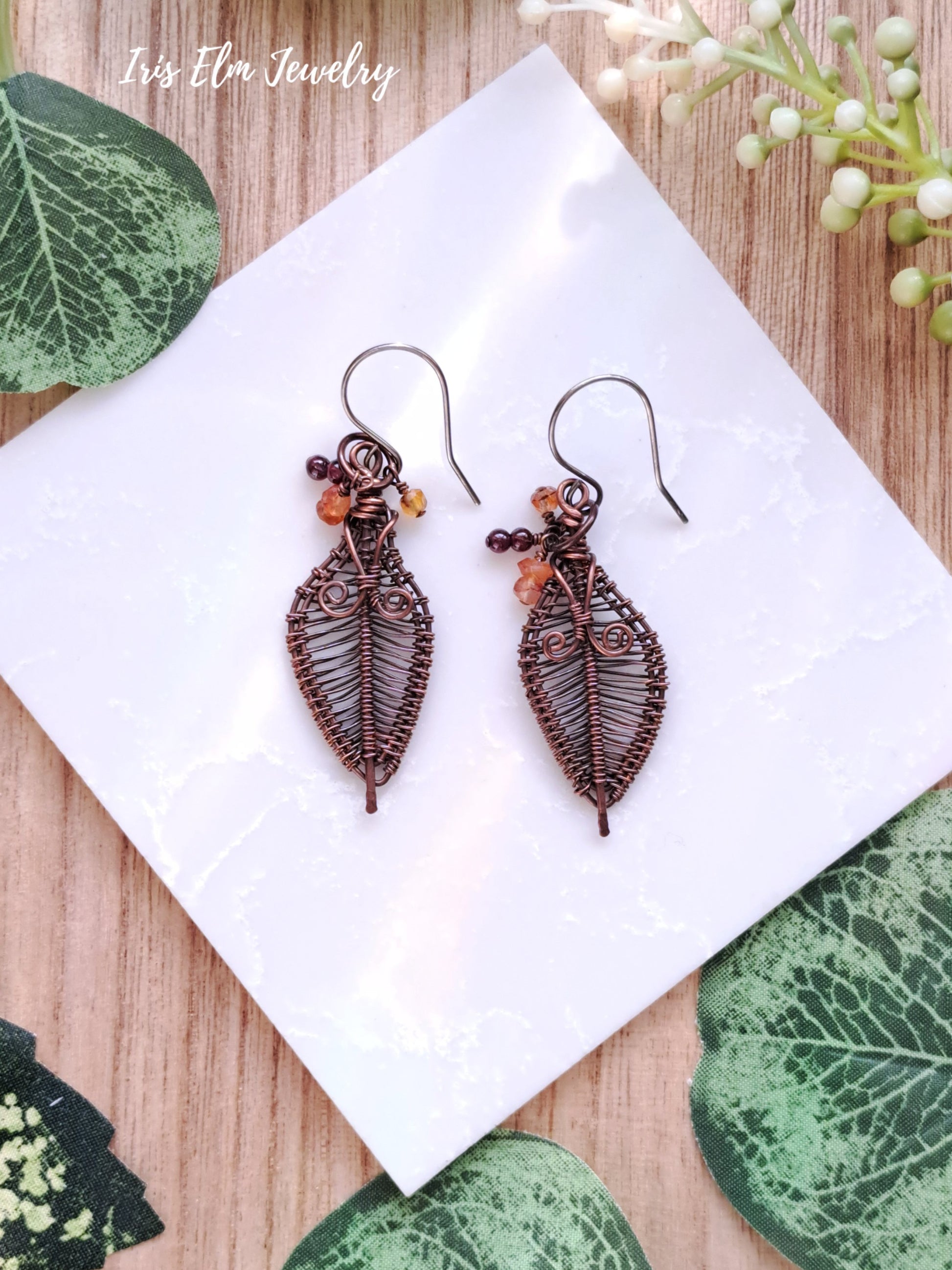 Pair of copper leaf-shaped earrings on a white tile with green leaves and flowers in the background.