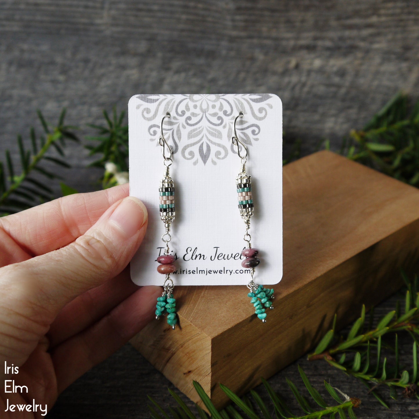 Pair of gemstone turquoise and rhodonite earrings on a 'Iris Elm Jewelry' card held by a hand with a natural background.
