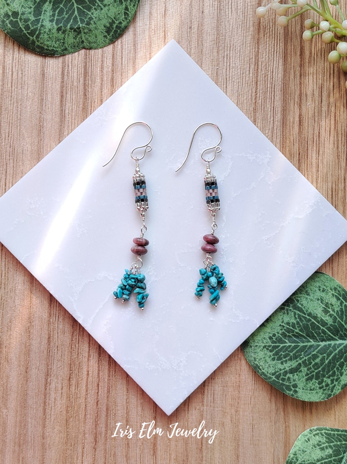 Pair of beaded turquoise and rhodonite stone and seed bead dangle earrings on a white card with green leaves in the background