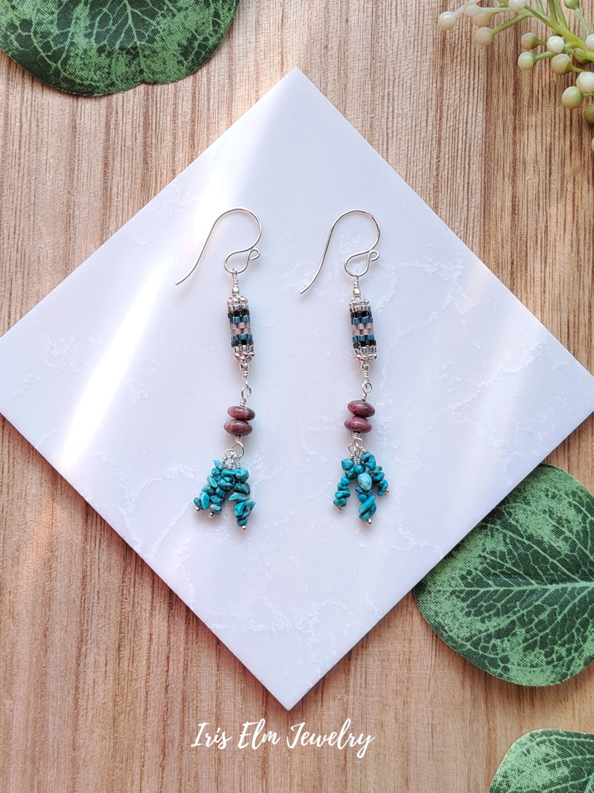 Pair of beaded turquoise and rhodonite stone and seed bead dangle earrings on a white card with green leaves in the background