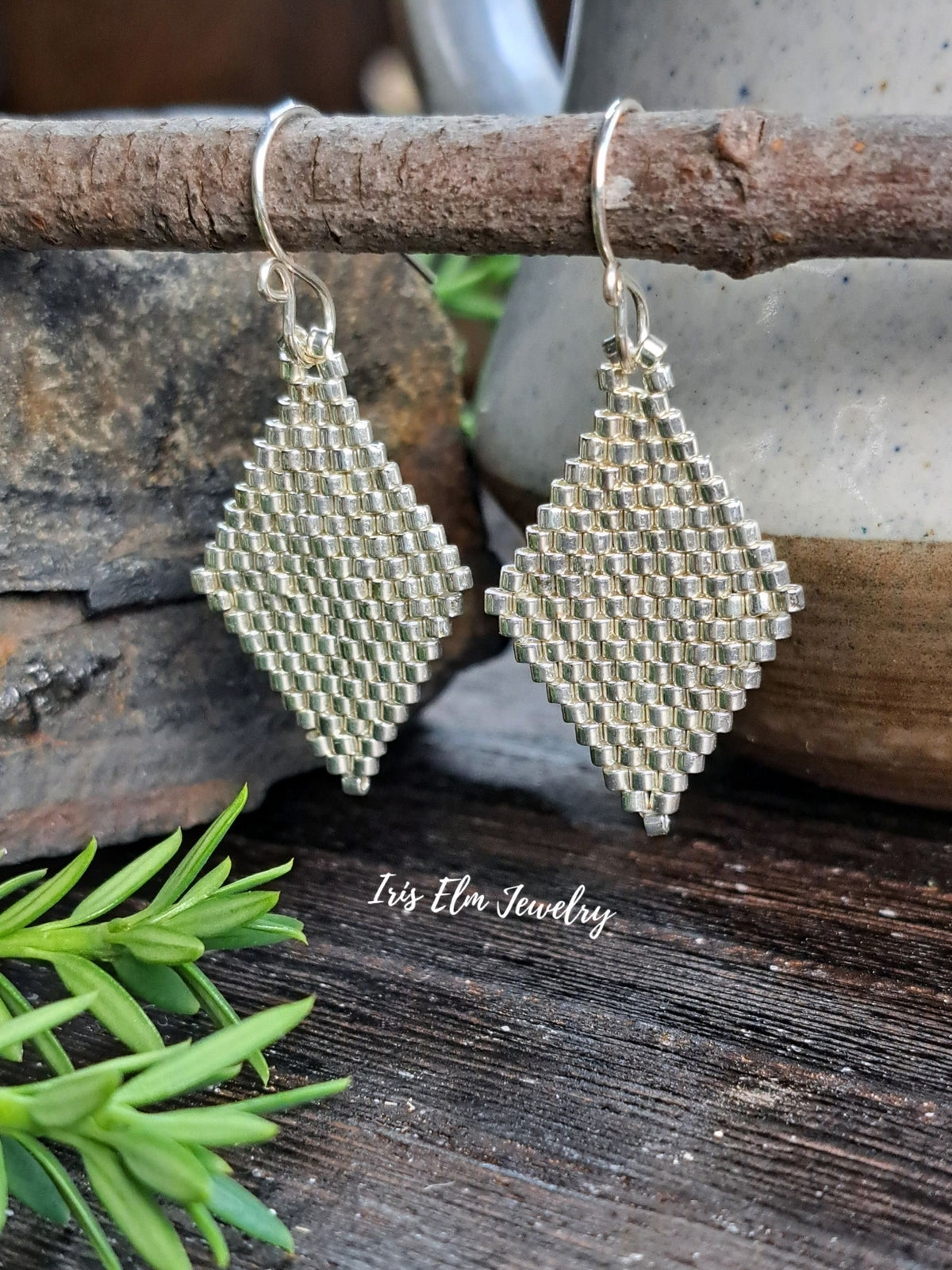 A pair of handmade beaded dangle earrings with diamond-shaped mesh bodies in a silver-tone metal, hanging from silver-tone fishhook wires. The earrings are displayed on a rustic wooden branch over a dark wood surface, with a ceramic pot and green sprigs in the background.