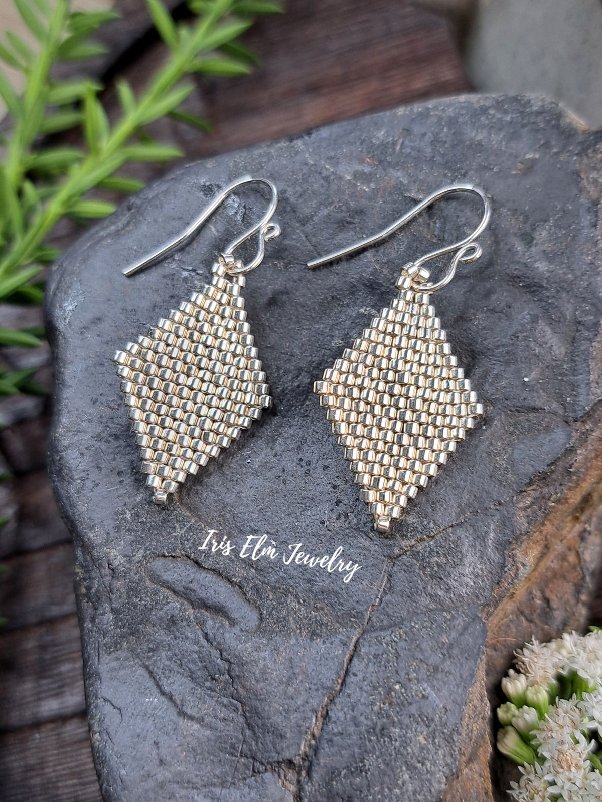 A pair of handmade silver seed bead drop earrings shaped like elongated diamonds, displayed on a dark textured stone with green leaves and small white flowers.