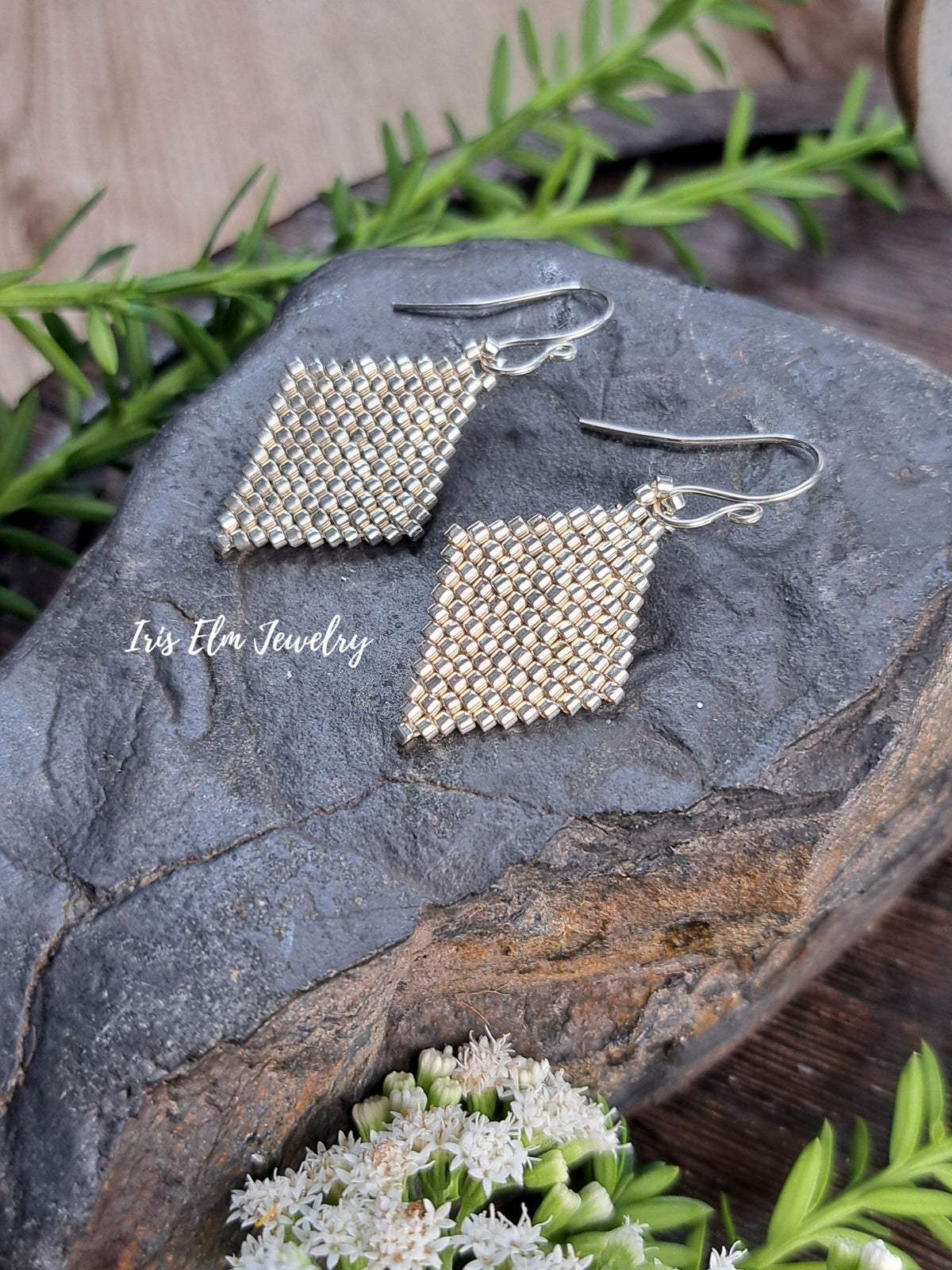 A pair of handmade square mesh drop earrings in silver and gold-tone metal, displayed on a dark textured stone with green foliage and small white flowers.