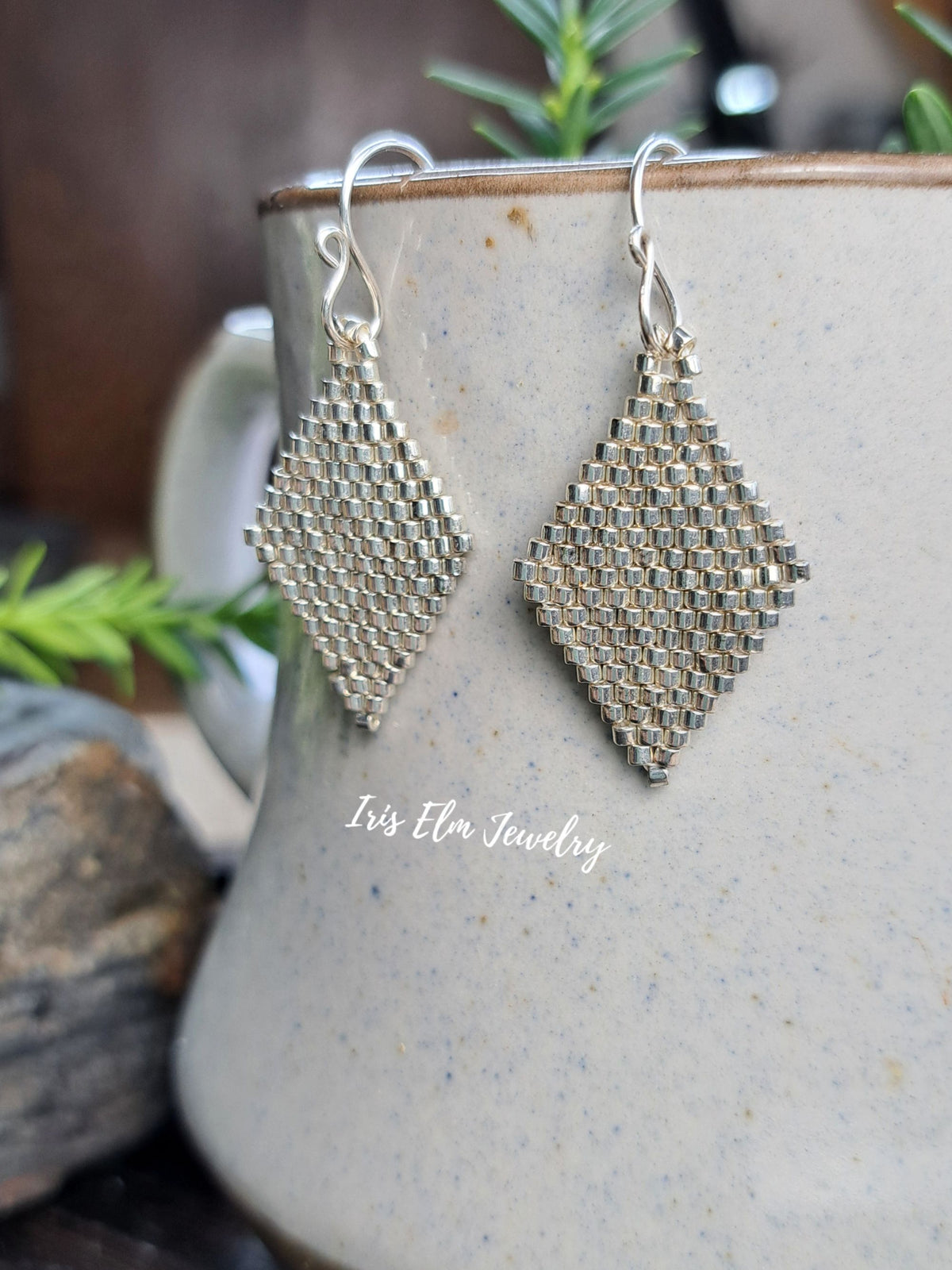 A pair of silver-tone seed bead handmade drop earrings with diamond-shaped dangling pendants, displayed in front of a speckled ceramic mug with a visible 'Iris Elm Jewelry' watermark.
