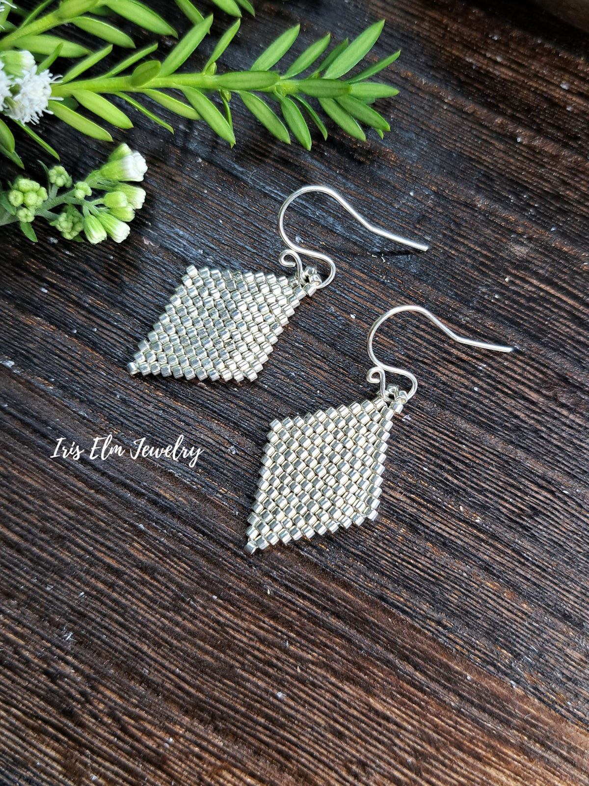 A pair of silver seed bead drop earrings with diamond-shaped pendants, displayed on a dark wood surface with green leaves and small white flowers. The brand name 'Iris Elm Jewelry' is watermarked on the left.