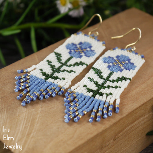 Beaded blue glass seed bead woven fringe earrings with floral folk art design on a wooden surface, featuring the brand 'Iris Elm Jewelry'.