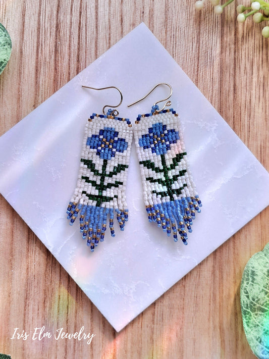 Beaded blue folk art flower seed bead fringe earrings on a white square with 'Iris Elm Jewelry' branding, placed on a wooden surface.