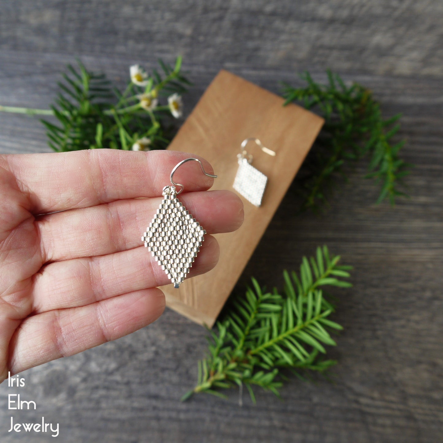 Silver geometric beadwoven seed bead earring held in a hand with a wooden display card and greenery in the background.