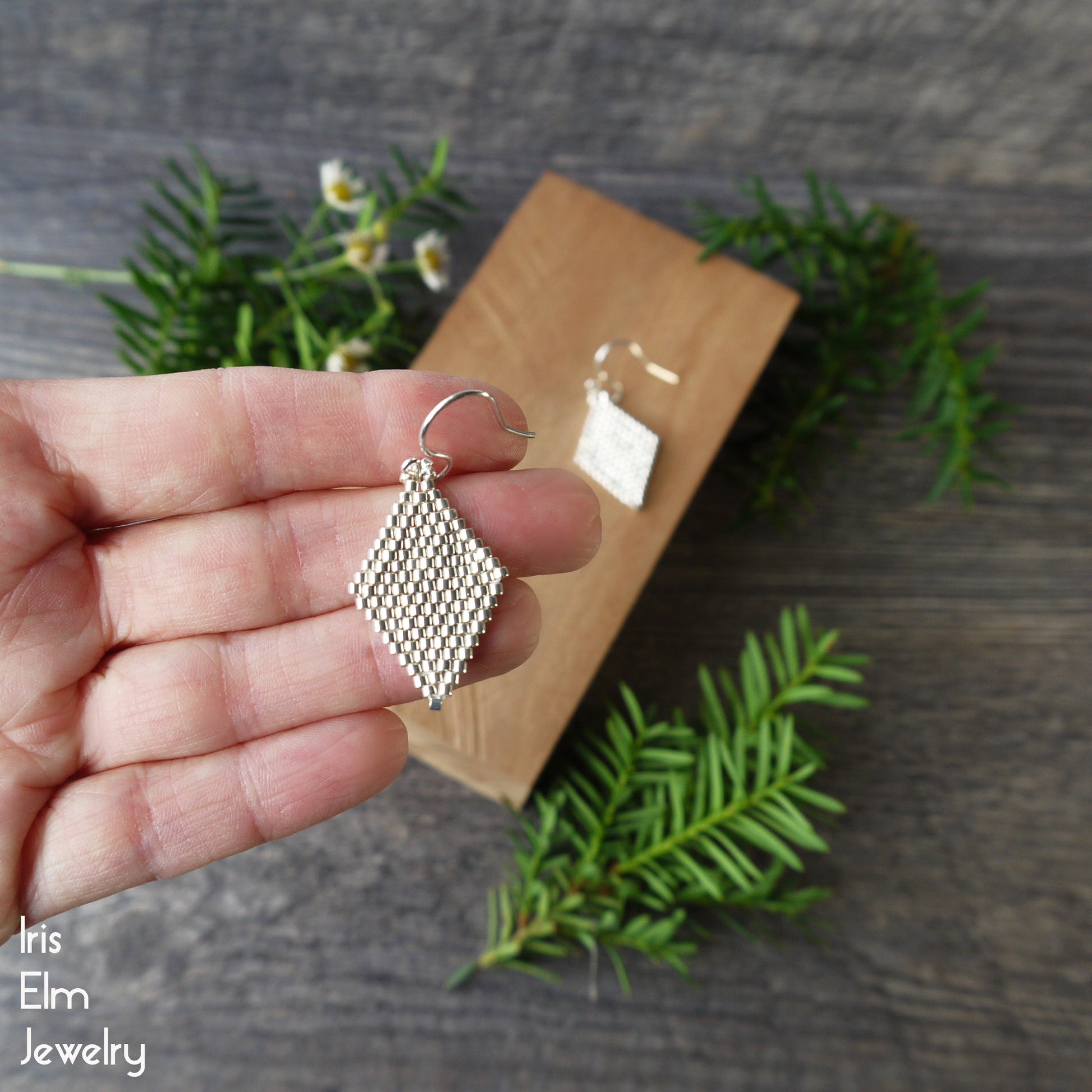 Silver geometric beadwoven seed bead earring held in a hand with a wooden display card and greenery in the background.