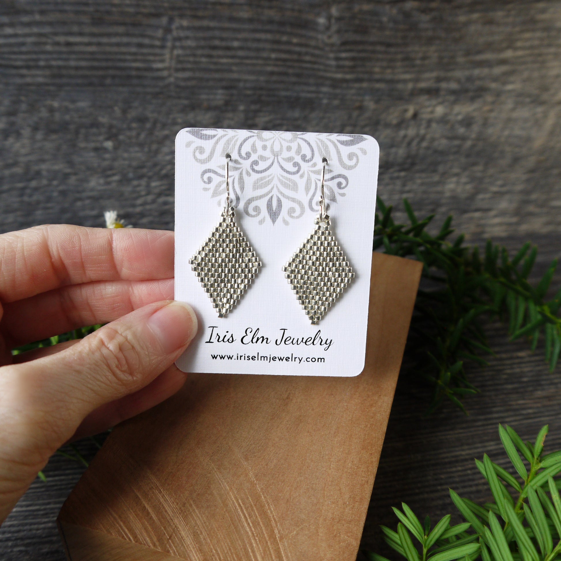 Pair of silver beaded earrings on a 'Iris Elm Jewelry' card held by a hand with a wooden surface and greenery in the background.