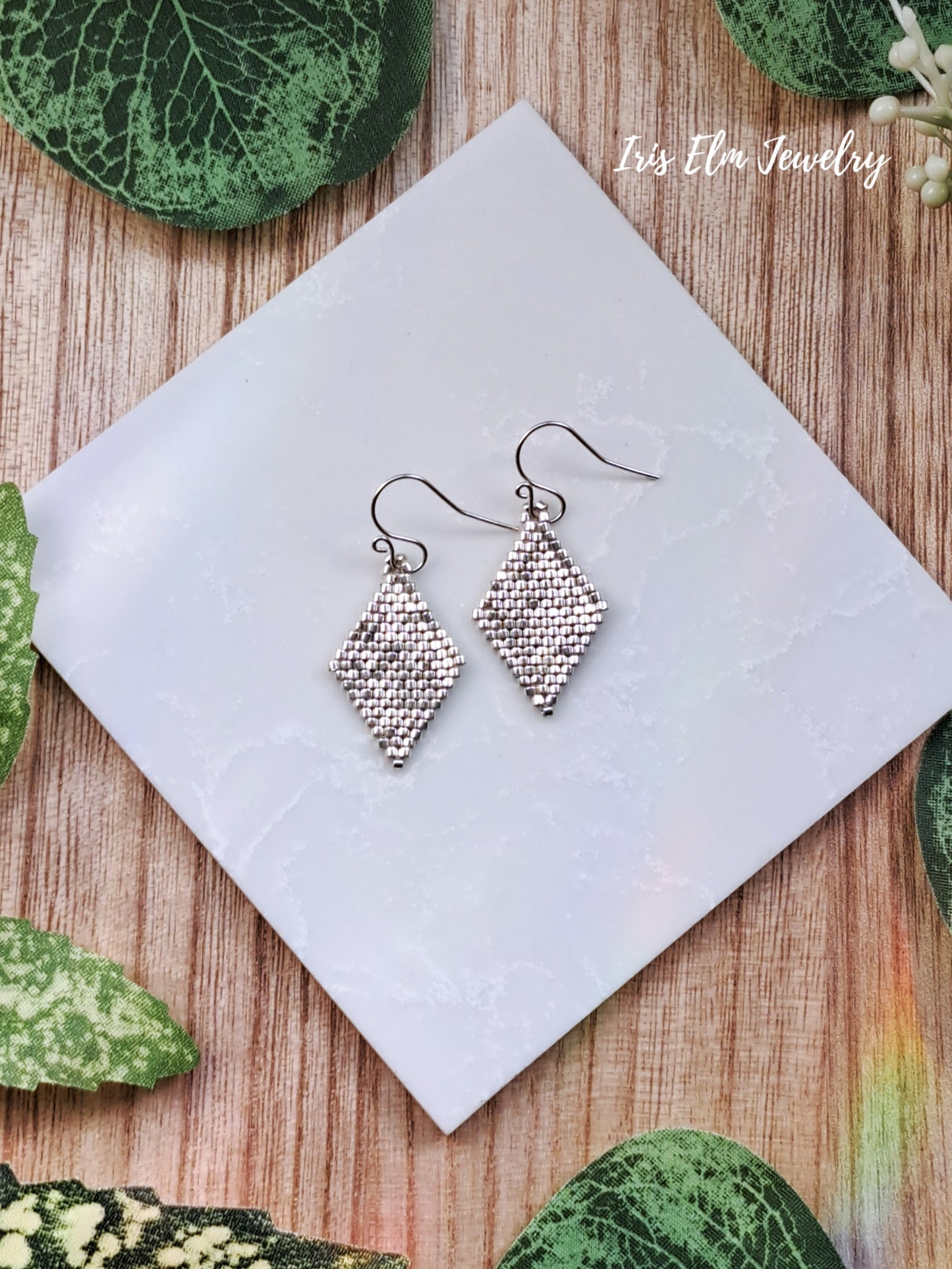 Silver glass seed bead woven diamond earrings on a white square with green leaves around
