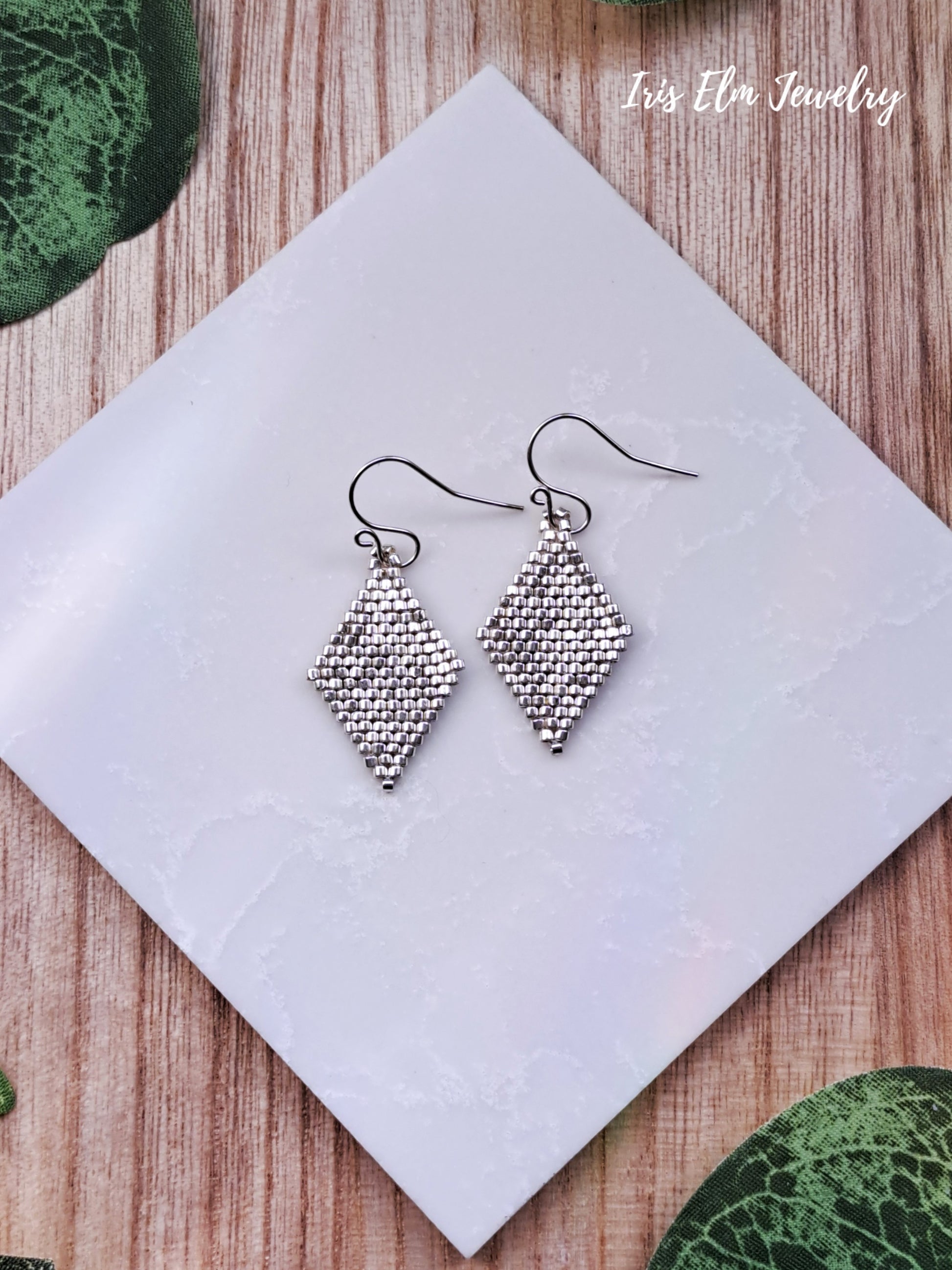 Silver seed bead diamond shape earrings on a white square card with 'Iris Elm Jewelry' branding, on a wooden surface.