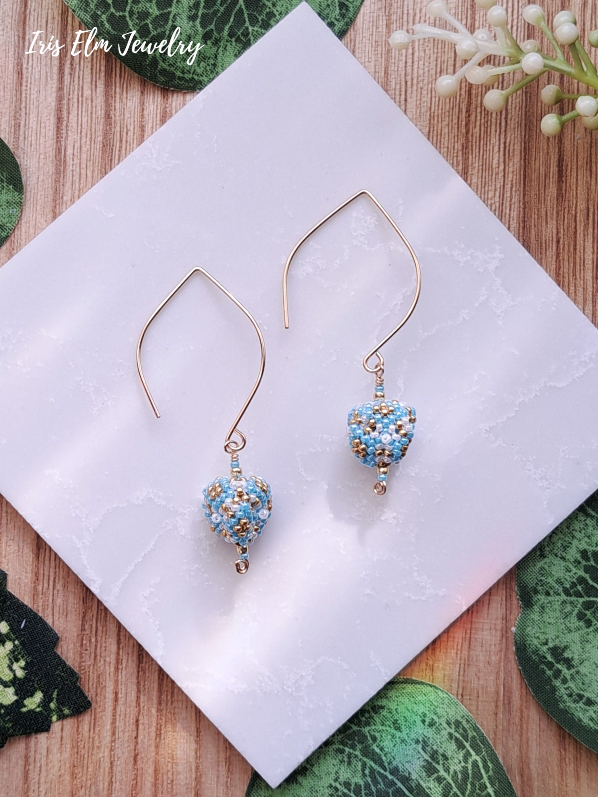 Pair of gold and light blue seed bead triangle beaded earrings on a white card with green leaves in the background