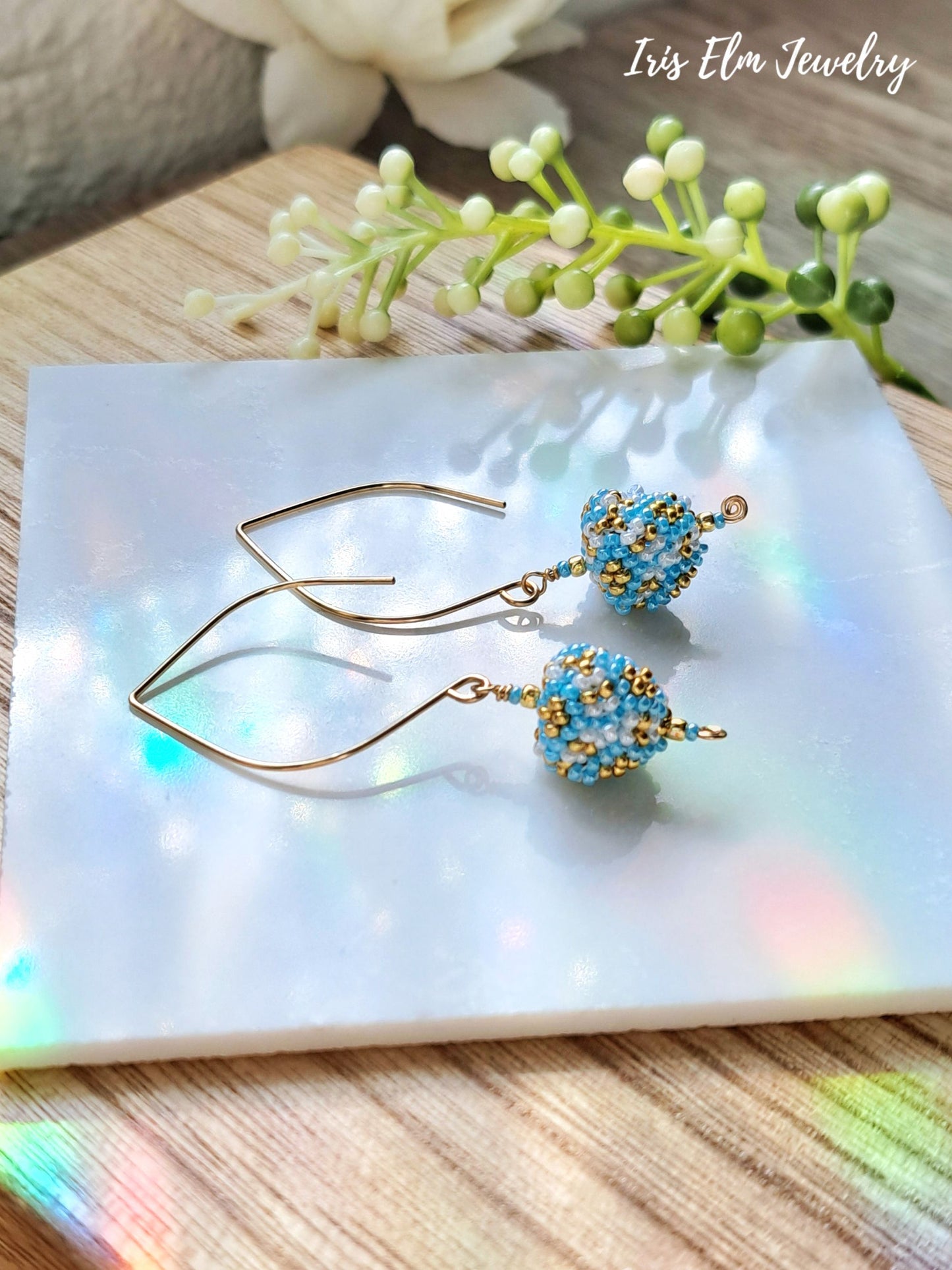 light blue, gold, white small seed bead triangle drop earrings on white marble surface with Iris Elm Jewelry branding.