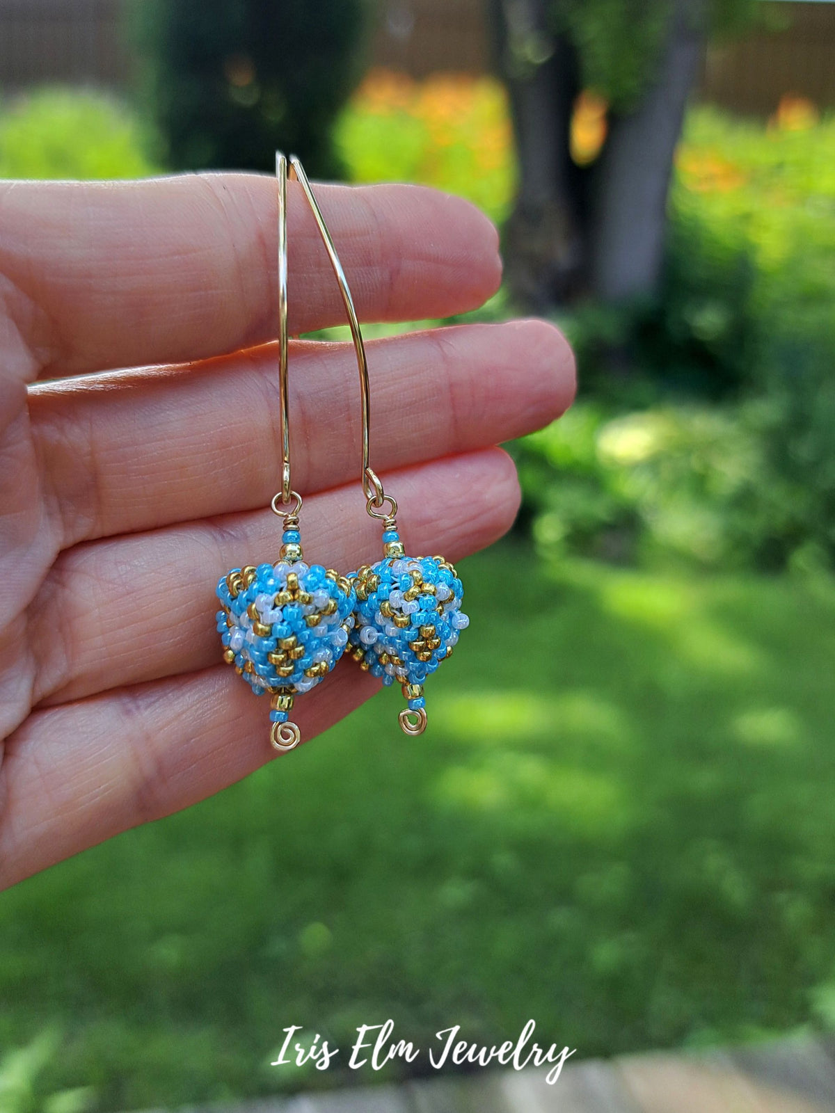 A hand holds a pair of handmade beaded drop earrings with gold wire hooks and blue, white, and gold seed bead heart pendants against a blurred green garden background. The text 'Iris Elm Jewelry' is visible at the bottom.