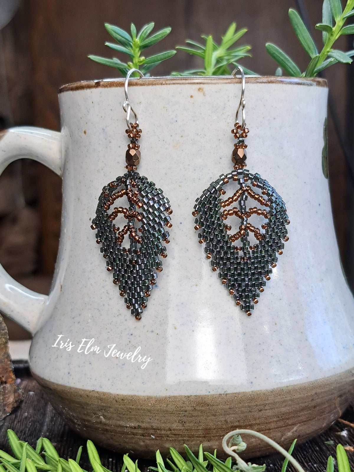 dark green and brown seed bead woven leaf earrings dangling from ceramic mug