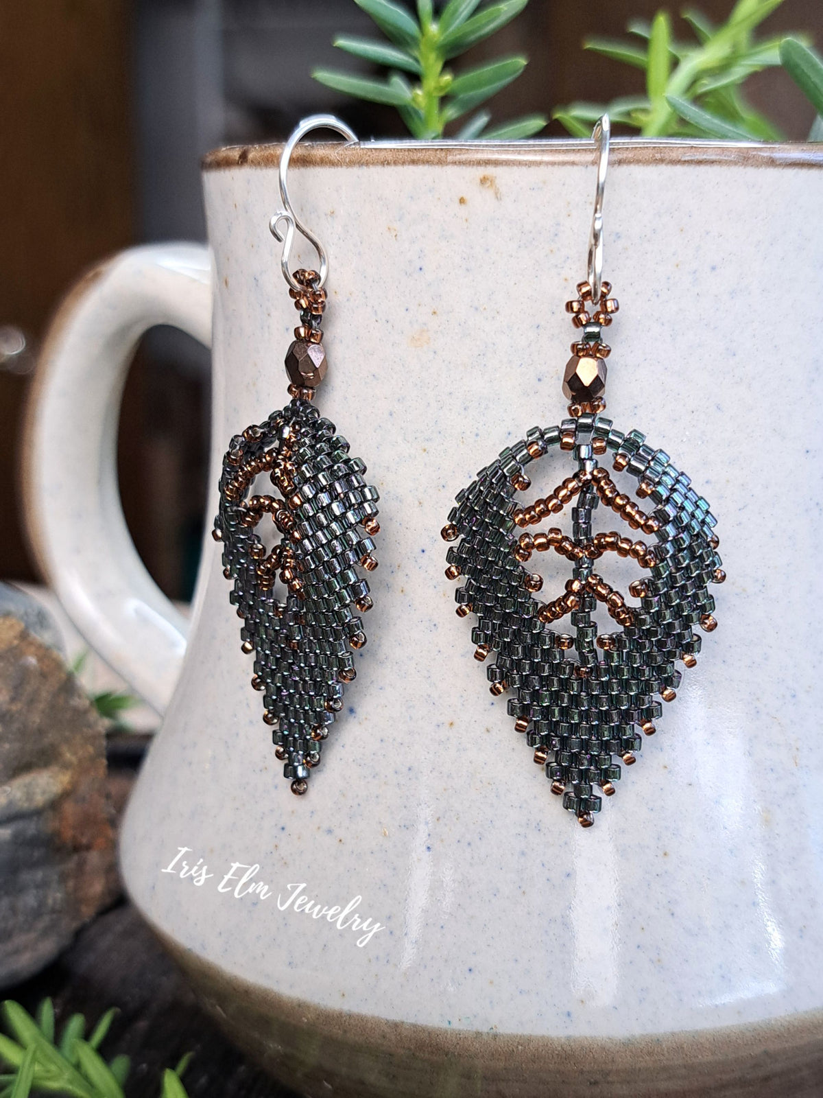 A pair of beaded leaf-shaped dangle earrings with dark gray and bronze seed beads, hanging in front of a ceramic mug. The earrings feature a geometric leaf design with a central branch motif.