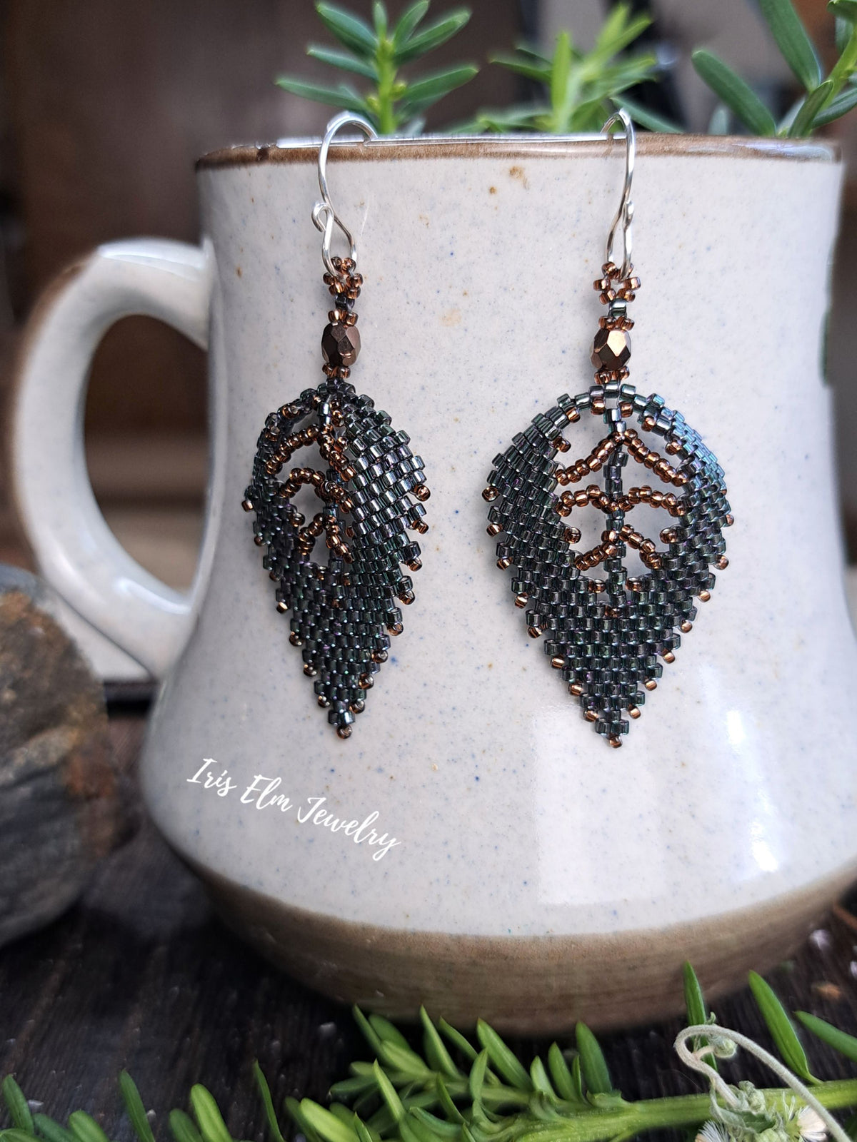 A pair of beaded dangle earrings shaped like leaves, featuring dark metallic beads and copper-toned wire detailing, hanging in front of a ceramic mug with green sprigs around it. The text 'Iris Elm Jewelry' is visible on the mug.
