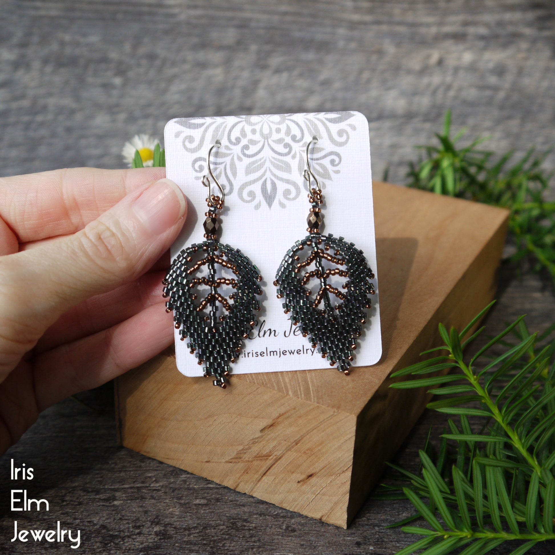 Pair of beaded brown and dark green leaf earrings on a 'Iris Elm Jewelry' card, held by a hand with a natural background.
