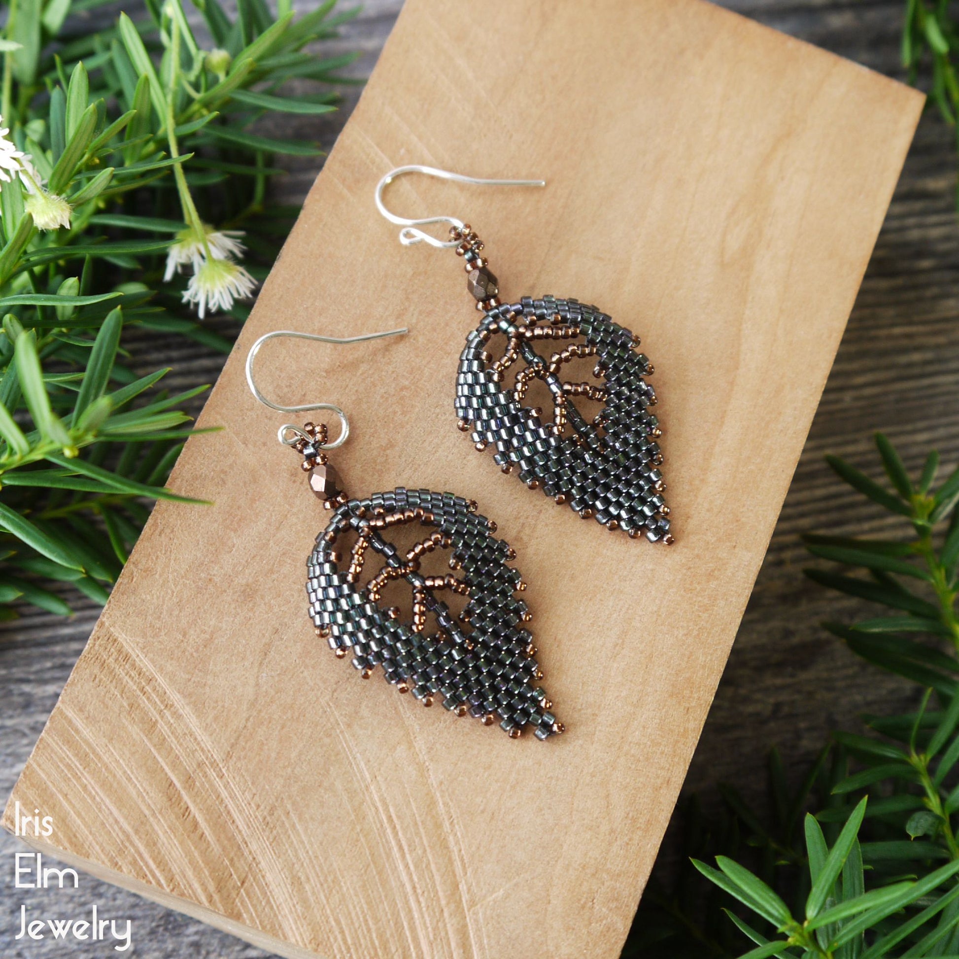 Pair of leaf-shaped woven seed bead green earrings on a wooden surface with greenery in the background