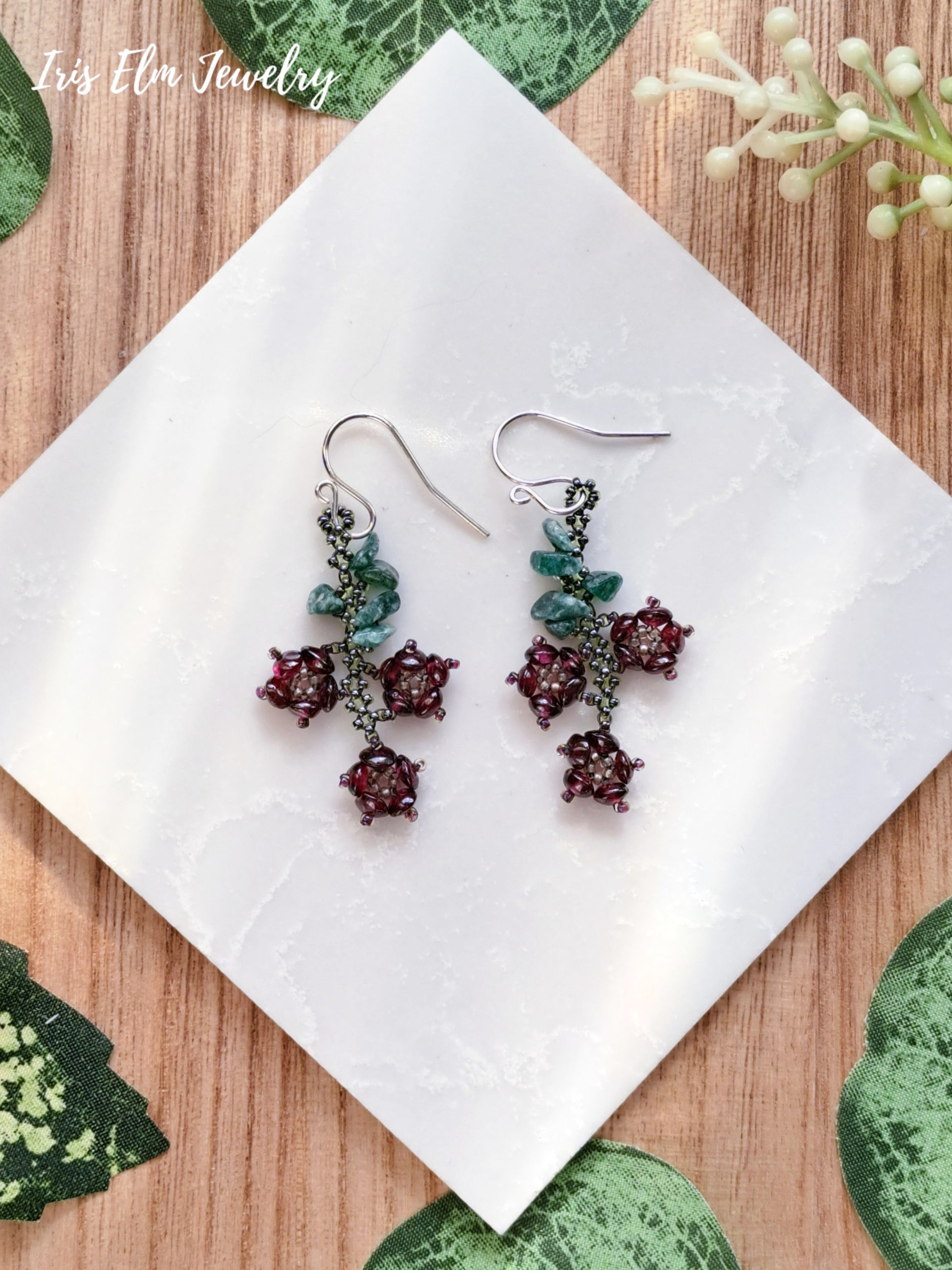 Pair of garnet and aventurine flower earrings on a white tile with green leaf patterns on a wooden surface.
