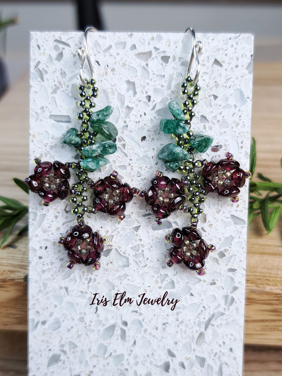 A pair of handmade beaded dangle earrings displayed on a white speckled card. The earrings feature green seed beads, clear faceted green crystals, and dark burgundy beads arranged in floral clusters.
