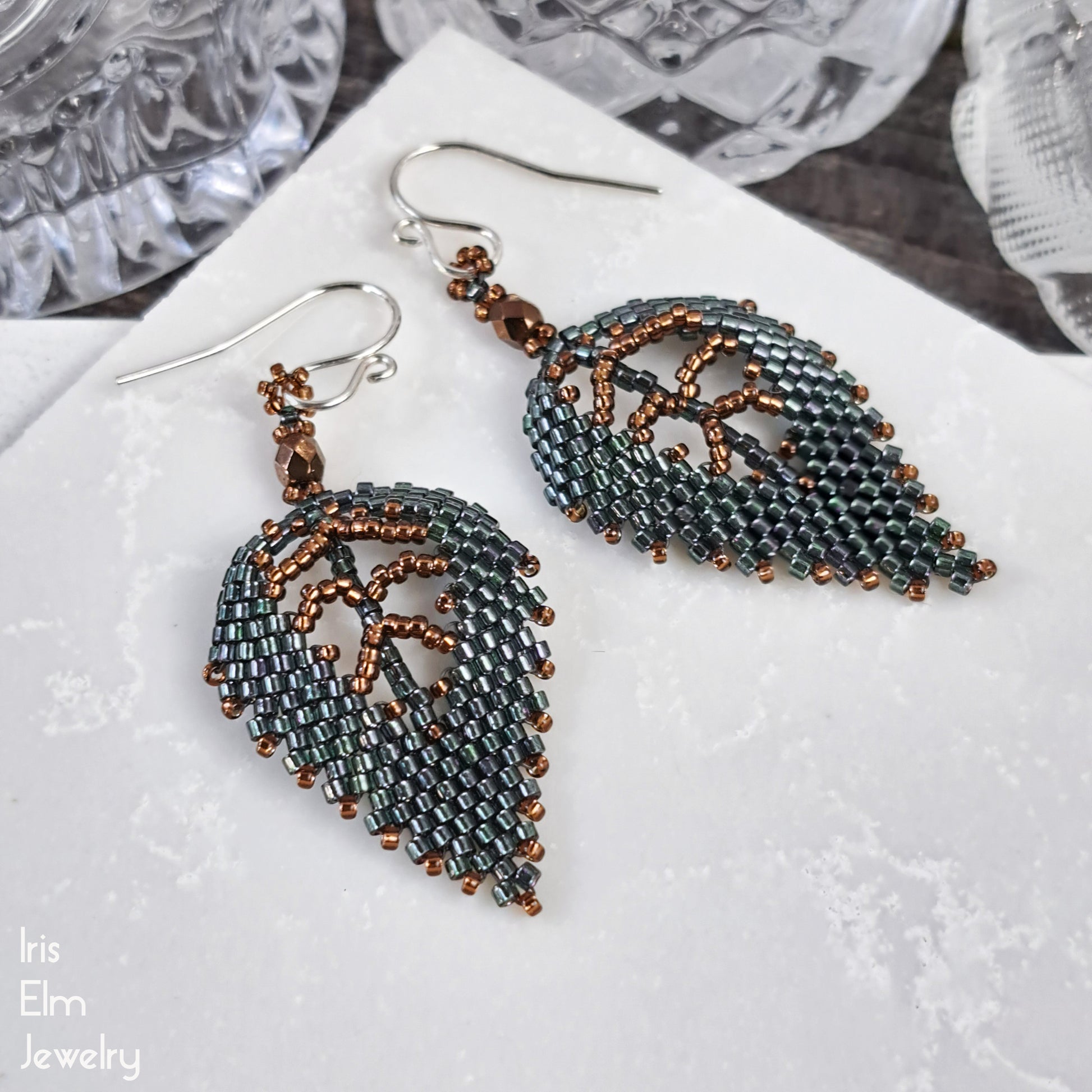 Pair of beaded dark green seed bead leaf earrings on a textured surface with 'Iris Elm Jewelry' branding.
