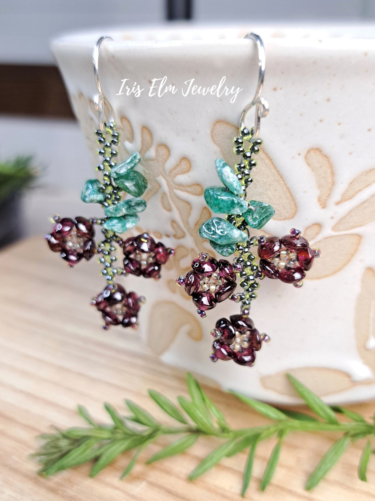 A pair of handmade beaded dangle earrings by Iris Elm Jewelry, featuring deep burgundy seed beads in flower clusters, light green beaded leaves, and teal-green rhinestone leaf accents, hanging in front of a ceramic mug on a wooden surface.