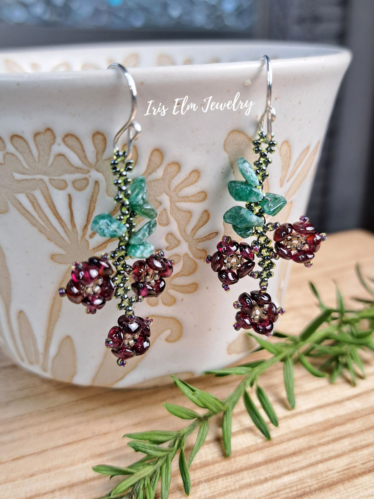 A pair of handmade beaded drop earrings by Iris Elm Jewelry, featuring deep burgundy berry clusters, green leaves, and a green beaded vine on silver hooks, displayed on a ceramic cup.