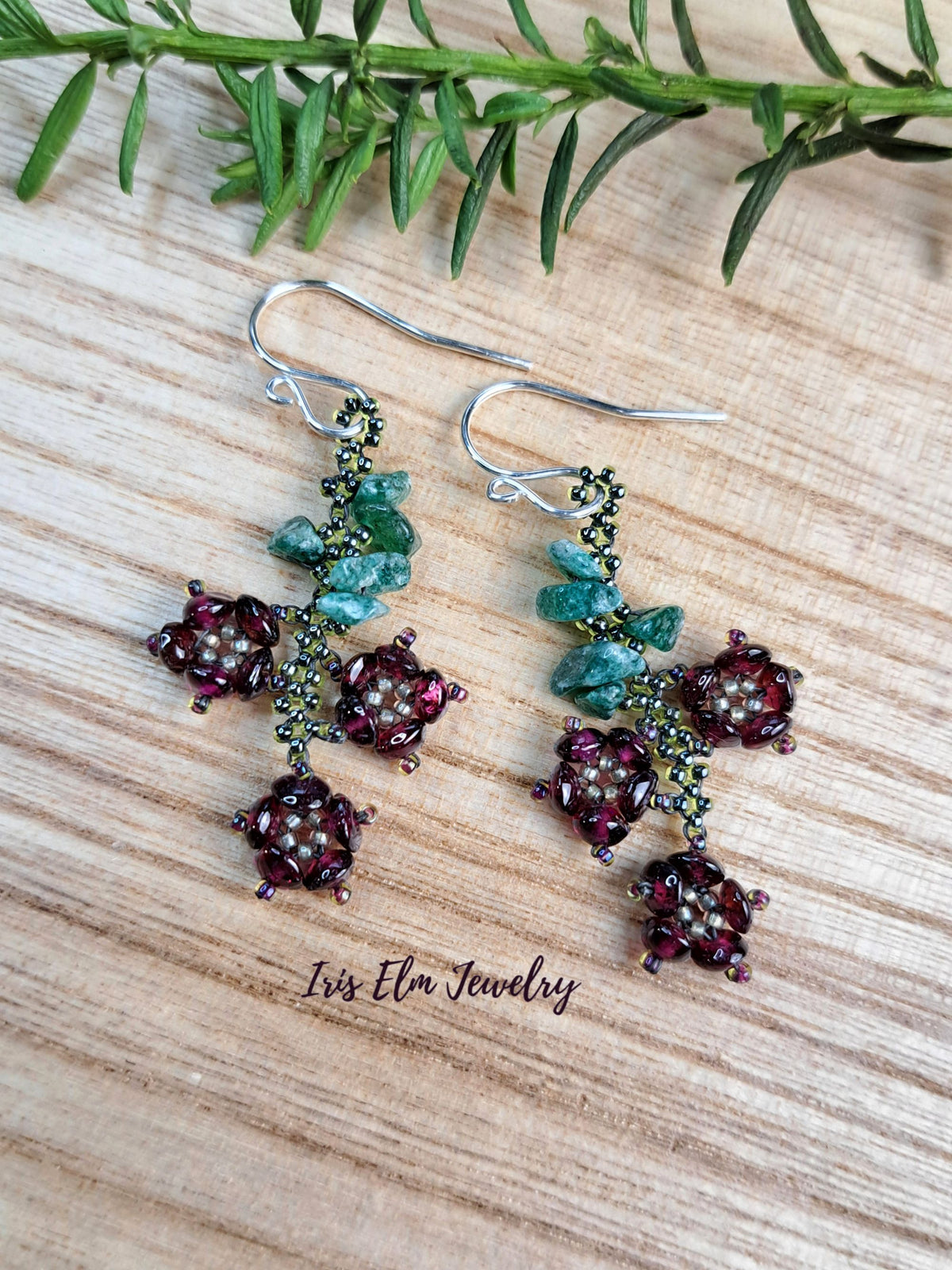 A pair of handmade beaded drop earrings with silver fishhook wires, featuring deep burgundy beads, green seed beads, and small turquoise-colored stone accents, displayed on a light wood surface with a sprig of green rosemary and the brand name 'Iris Elm Jewelry' printed below.