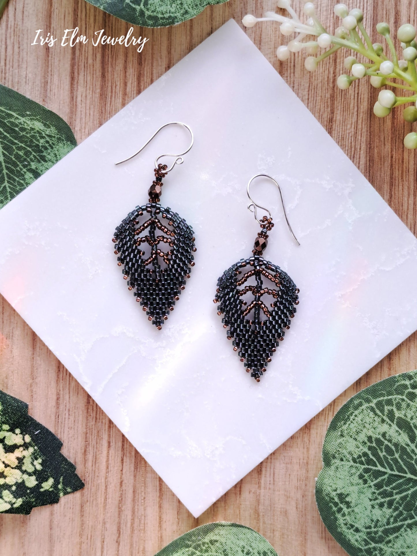 dark green seed bead leaf-shaped earrings on a white card with green leaves in the background