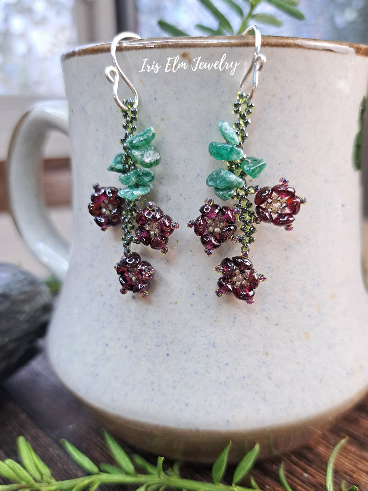 A pair of dangling earrings displayed on a speckled ceramic mug. The earrings feature green faceted stone leaves and clusters of deep burgundy-red flower-like beads on silver hooks.