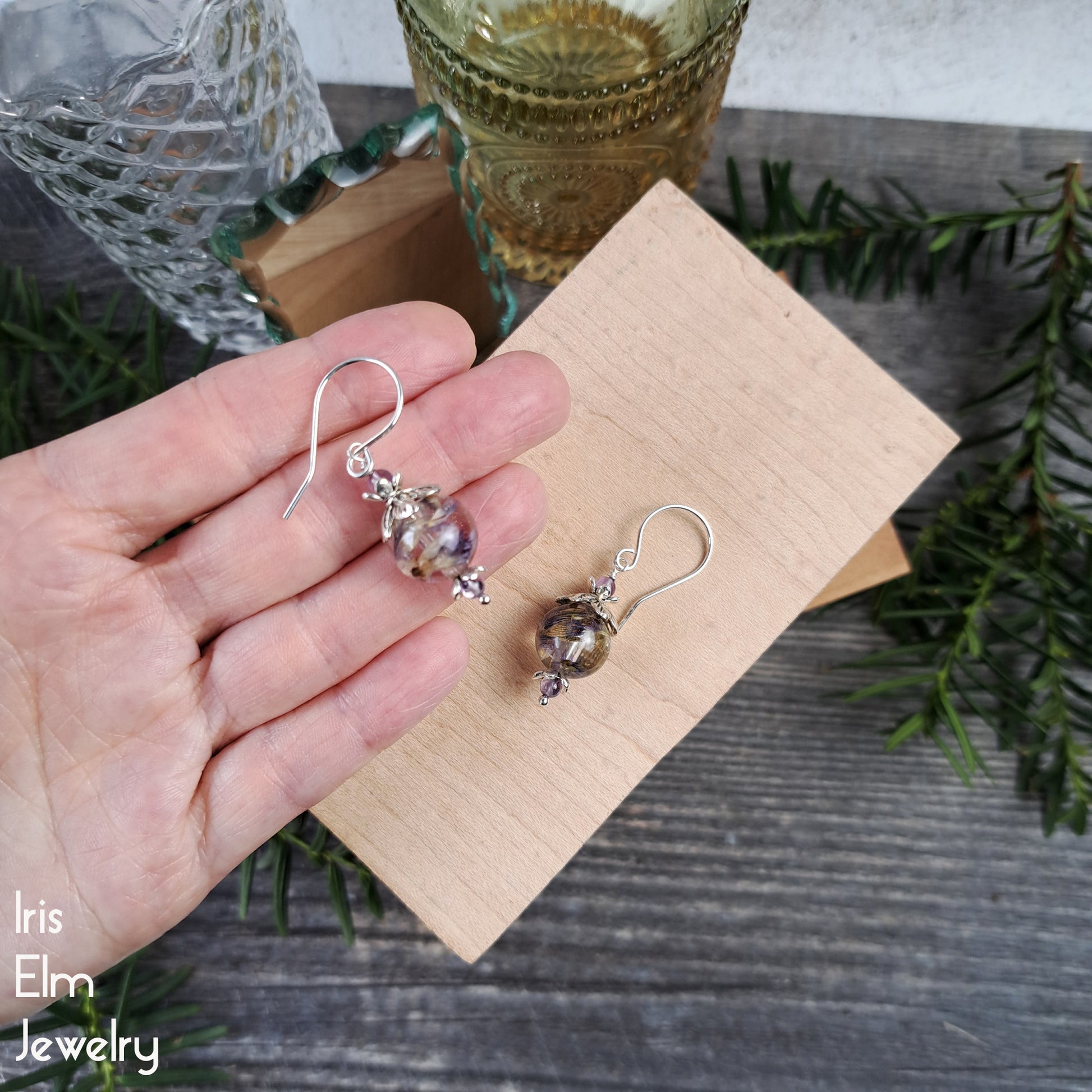 Hand holding a pair of silver earrings with purple flowers in resin beads and sterling silver earwires on a wooden surface.