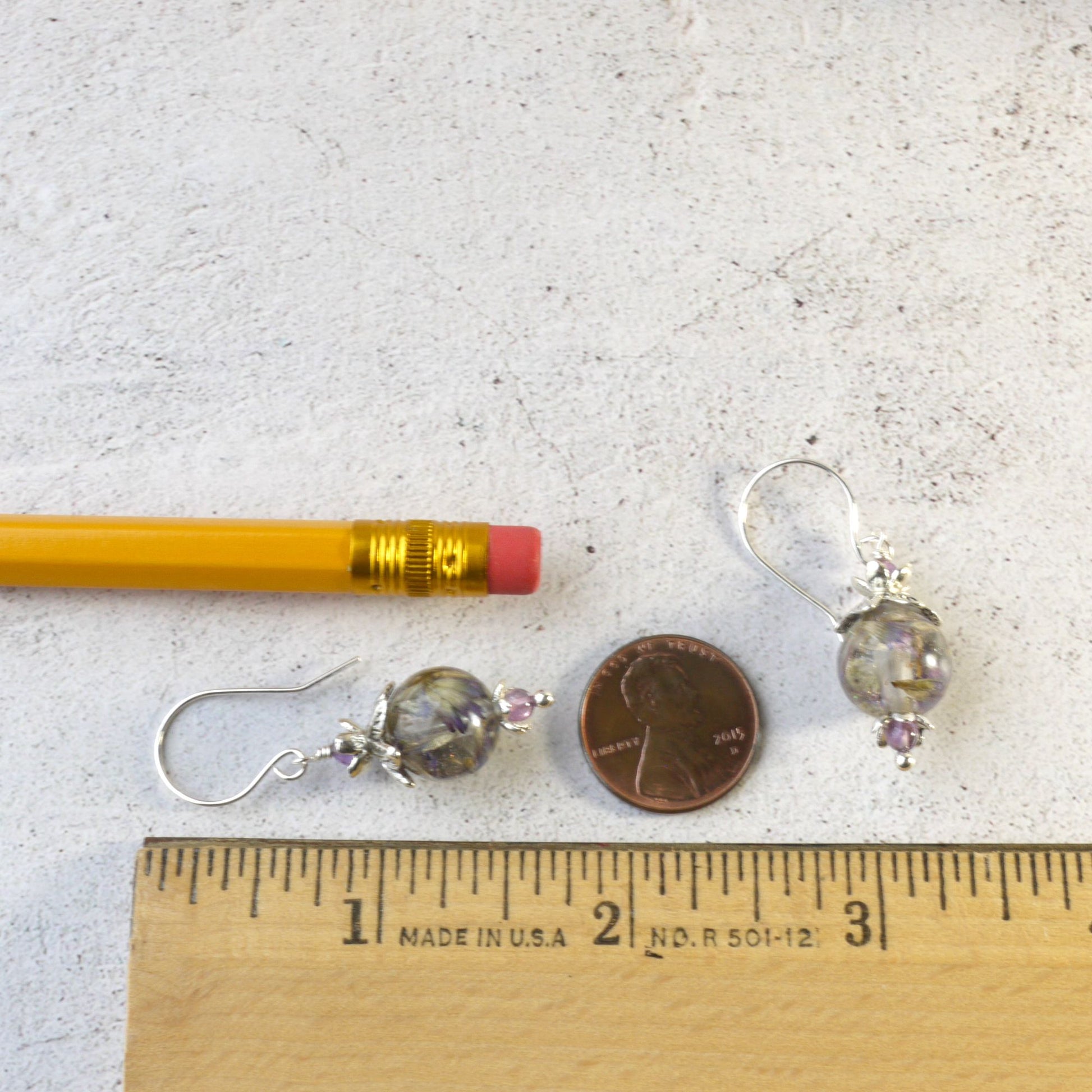 Pair of 1 and 5/8 inch long earrings with clear resin with purple flowers and amethyst beads next to a penny and ruler for size comparison on a light gray background. 