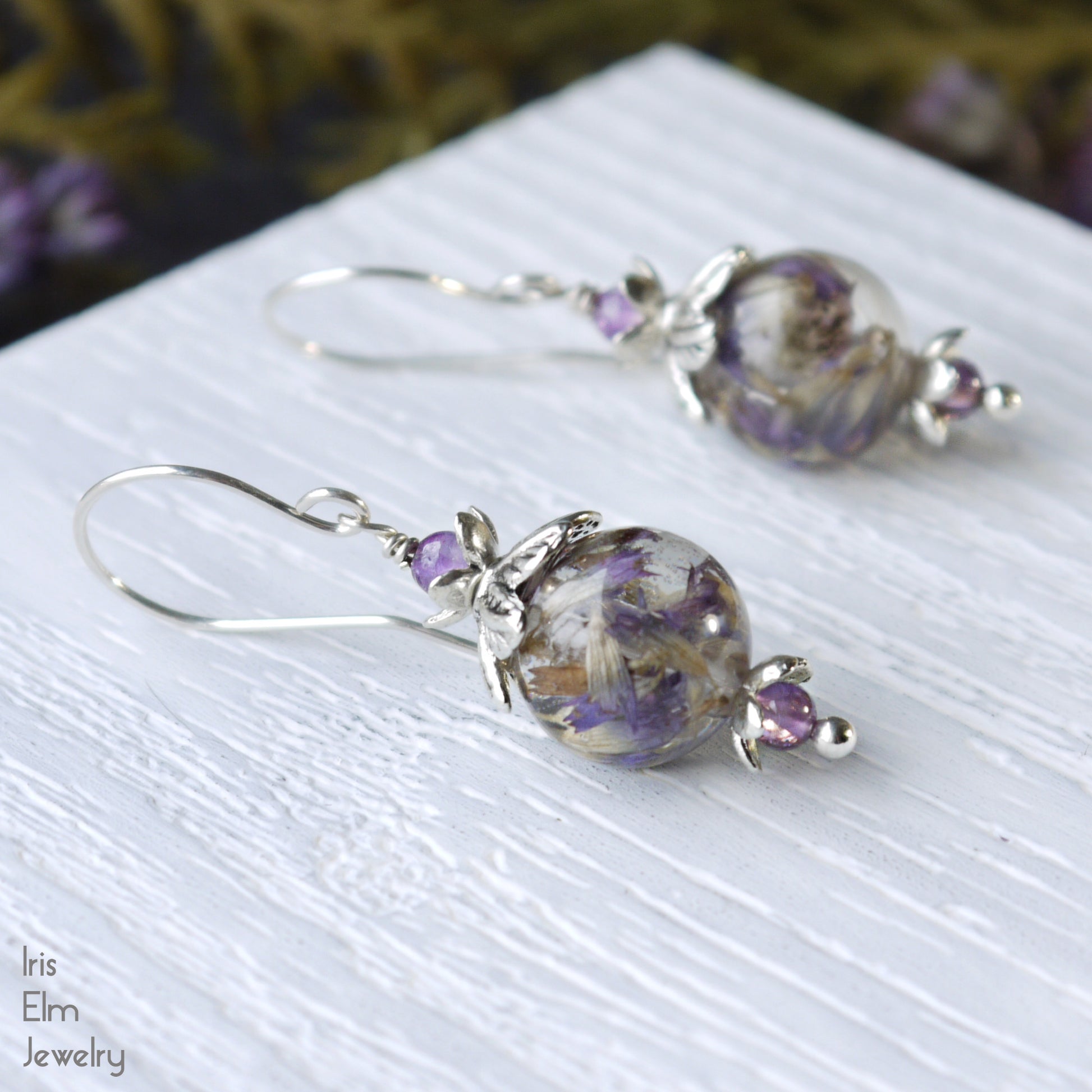 Silver earrings with purple flowers in resin and amethyst stone beads on a white surface, branded 'Iris Elm Jewelry'.